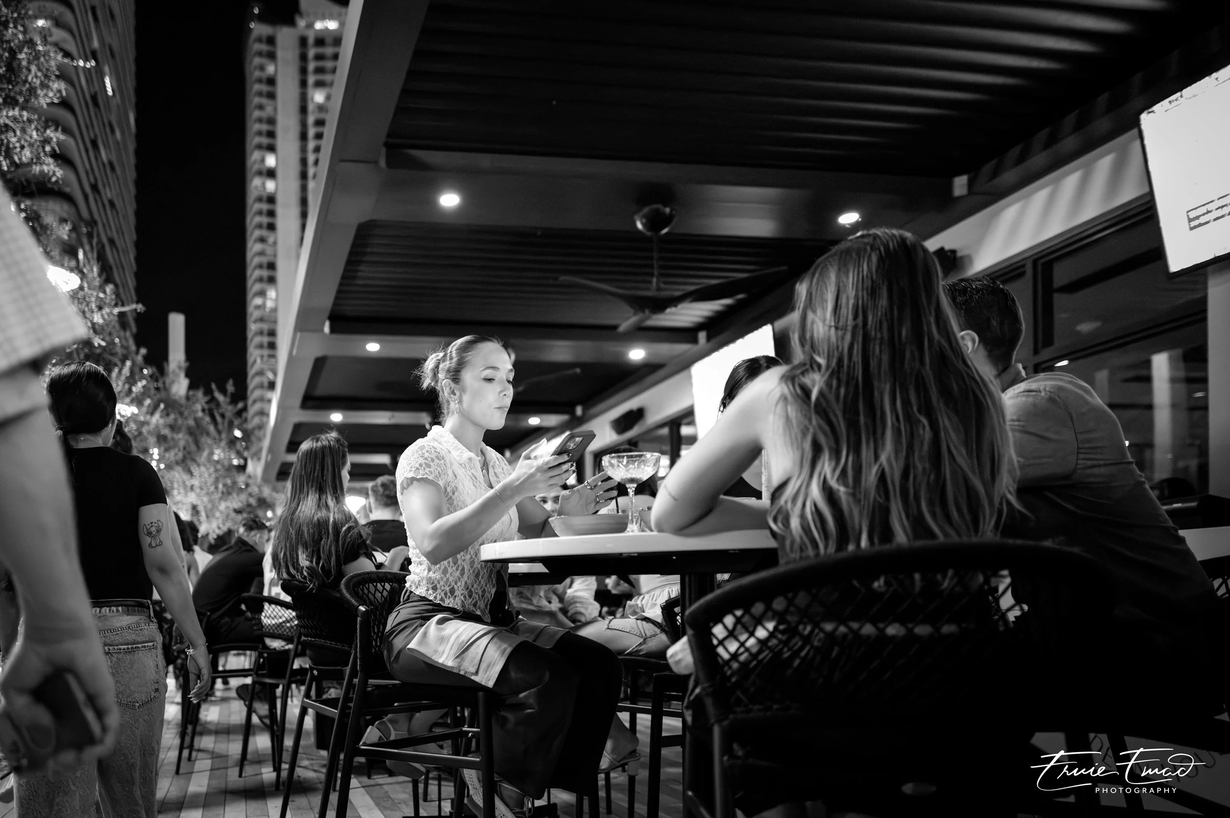Nighttime scene in a restaurant or bar with several people sitting at tables, one young woman reading a menu or phone, others engaged in conversation, decorated with string lights and a Christmas tree, modern architecture in the background.