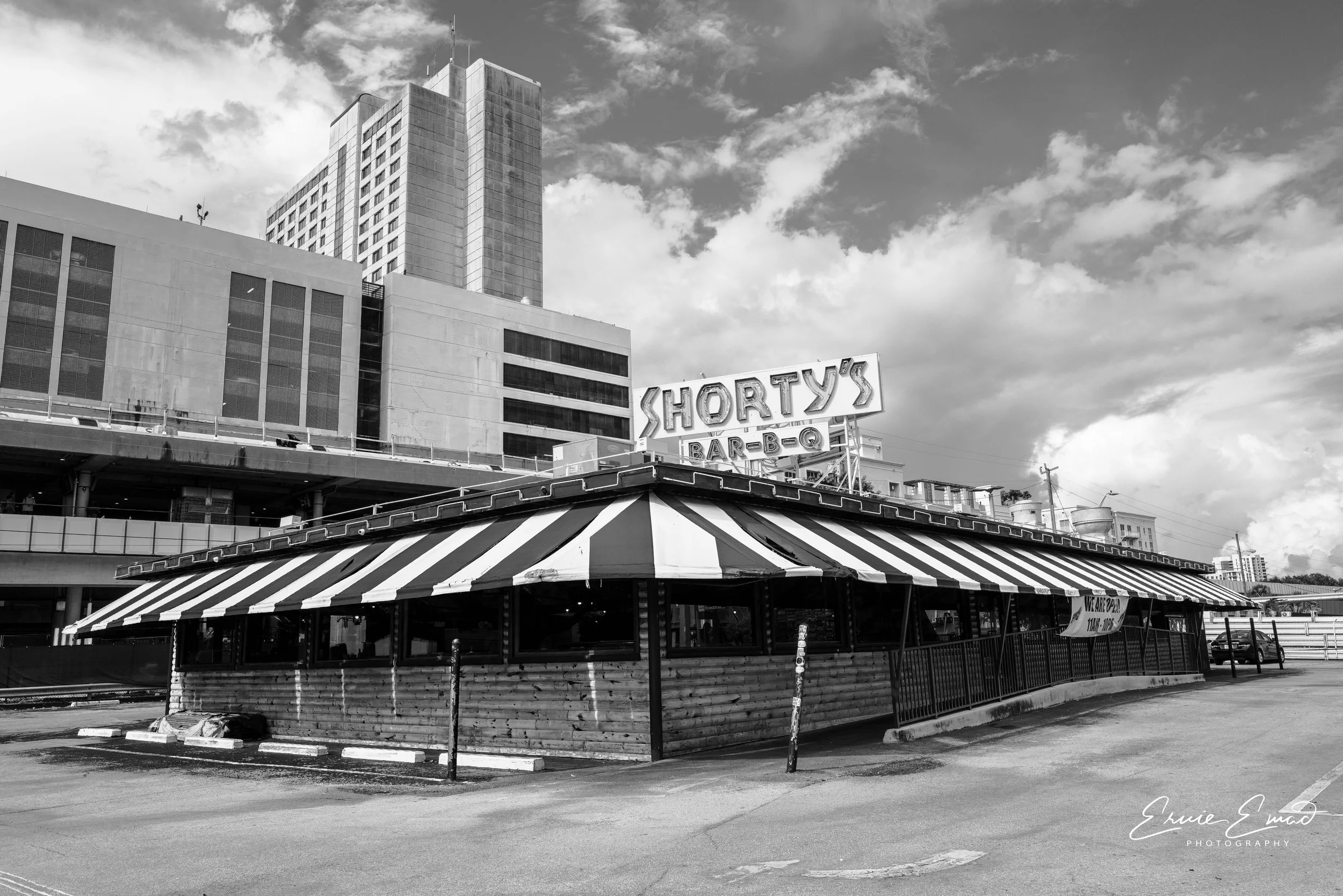 A black and white photo of a cityscape showing a large multi-story building with a retail or restaurant sign on top that reads 'Shorty's Bar-B-Q'. In the foreground, there is a stand with a striped roof, likely a food stall or closed outdoor seating area. The sky has some scattered clouds.