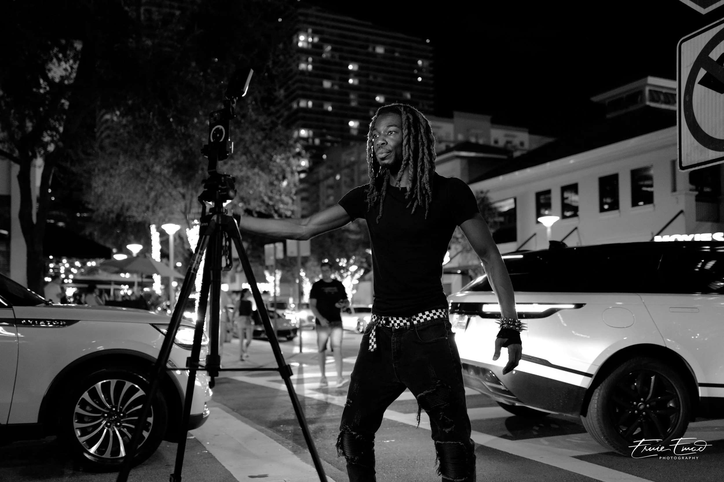 A person with dreadlocks and ripped jeans operating a camera on a tripod on a city street at night, illuminated by streetlights and building lights.