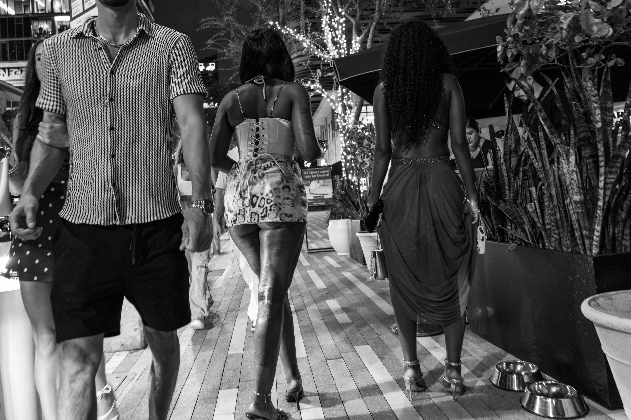 Night scene on a city sidewalk with several women and one man walking past a vendor stand. The women are dressed in trendy, revealing clothing with one wearing a corset and patterned shorts, and all with curly or wavy hair. Some are holding shopping bags or carrying items. The vendor stand has potted plants and metal pet bowls in front. Behind, there are string lights on trees and illuminated shop windows.