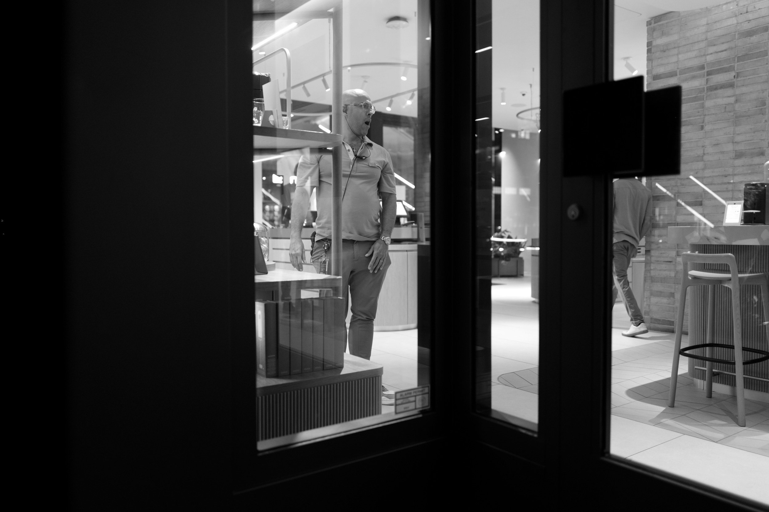 A black and white photo of a man inside a store, viewed through a glass door. The man is bald, wearing glasses, a short-sleeved polo shirt, and pants. He has earbuds in his ears and is looking to his left with a surprised or shocked expression. The s