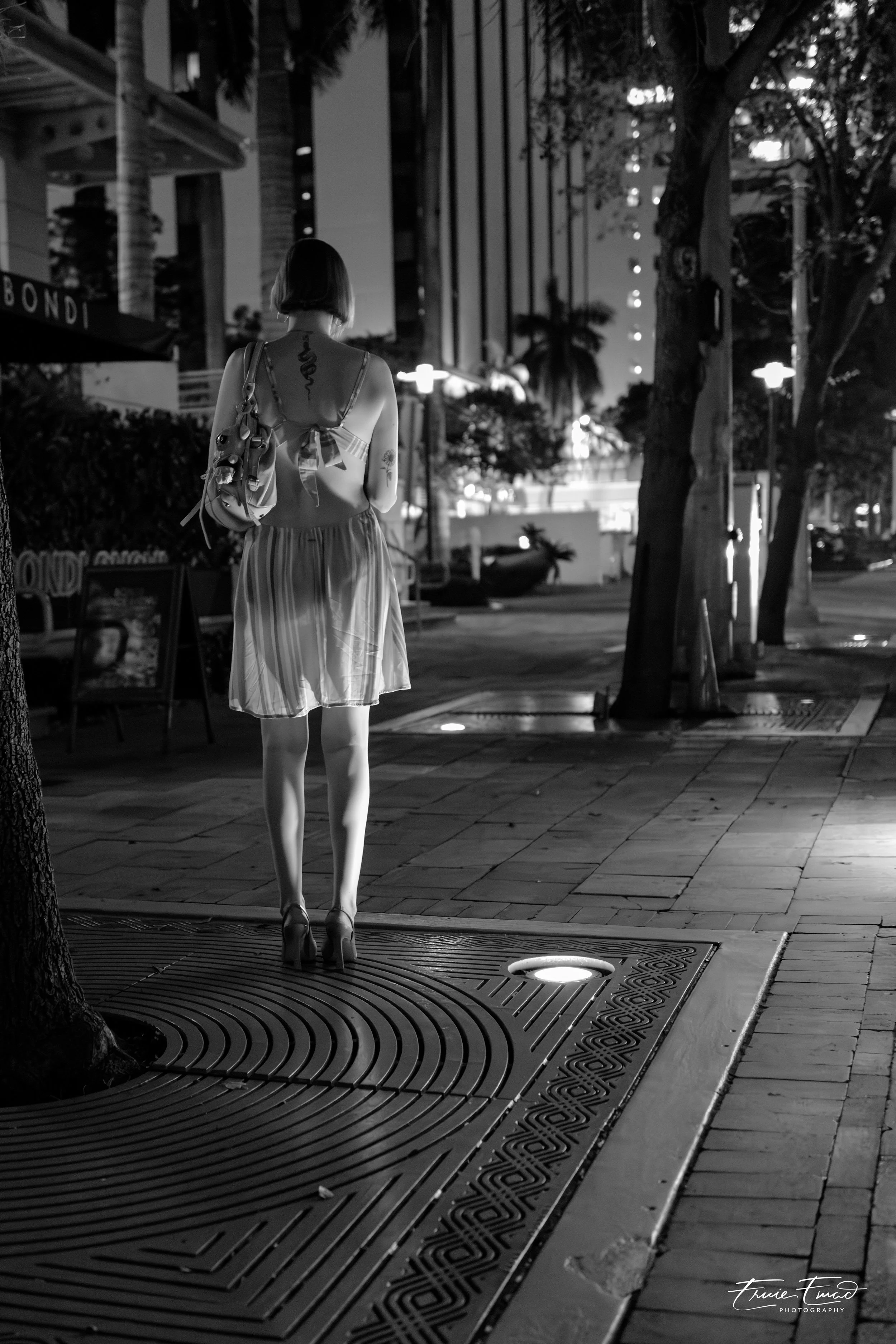 A woman with short hair wearing a striped dress and high heels walking alone on a city sidewalk at night, carrying a tote bag with sunglasses clipped to it, surrounded by streetlights and urban buildings, in black and white.