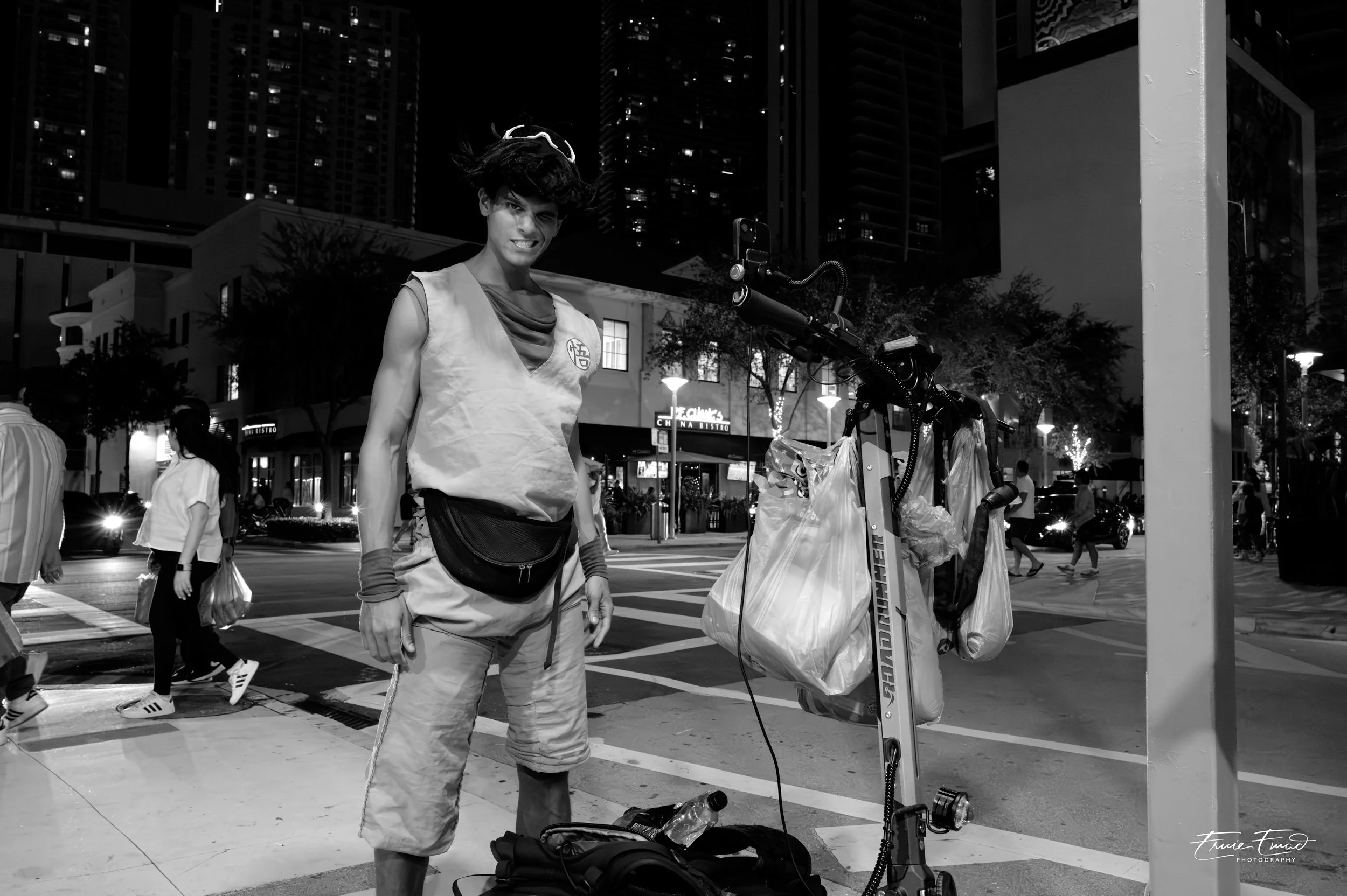 A street performer dressed in traditional Chinese attire standing next to a scooter with a large mechanical flower on the street at night, with tall buildings and city lights in the background.