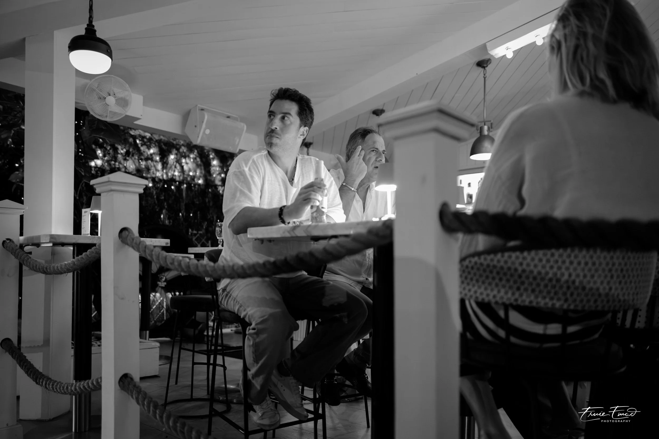 Black and white photo of three people sitting at a table inside a restaurant, with a woman in the foreground and two men behind her, engaged in conversation.