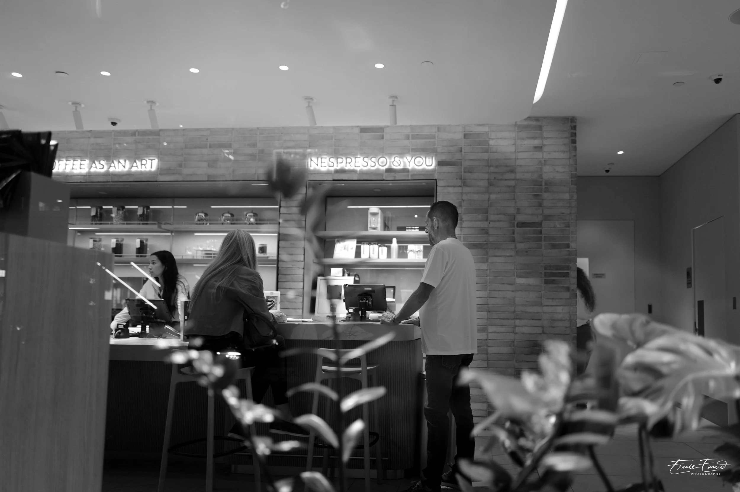 Inside a coffee shop with a brushed brick wall, neon signs reading "Coffee as an Art" and "Nespresso & You," and customers being served by staff members. The image is in black and white.