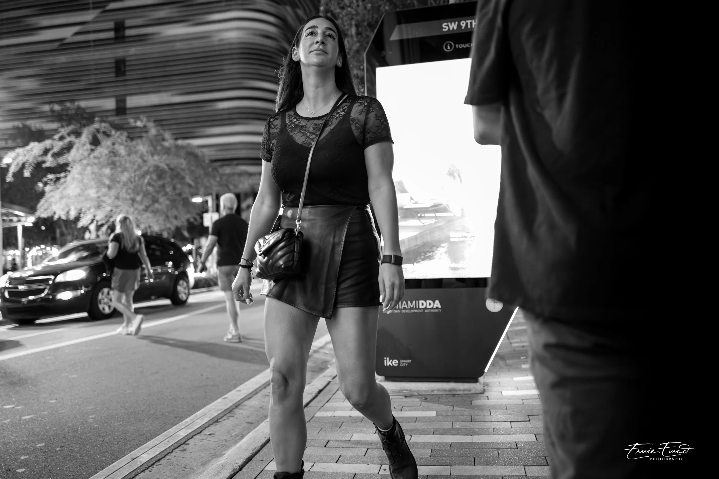 A woman in a leather skirt and lace top walking on a city sidewalk near a bus stop, with cars on the street and other pedestrians in the background, in black and white.