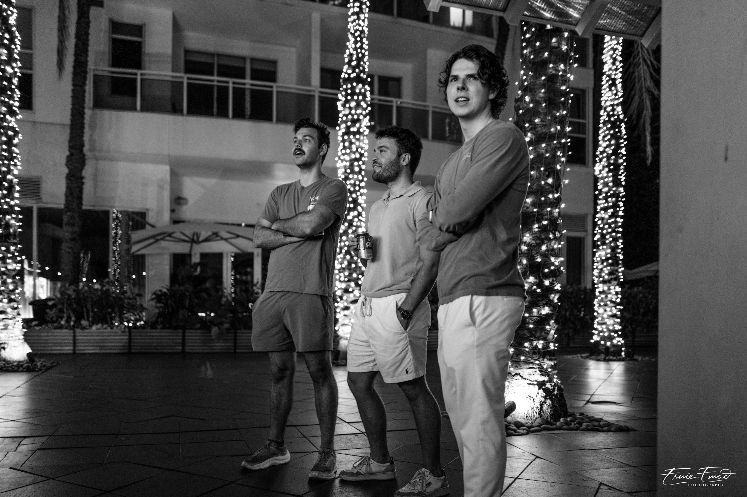 Three men standing outdoors at night near palm trees wrapped with string lights. Two have their arms crossed, one is holding a drink, and they appear to be engaged in conversation or watching something. Residential buildings are visible in the backgr