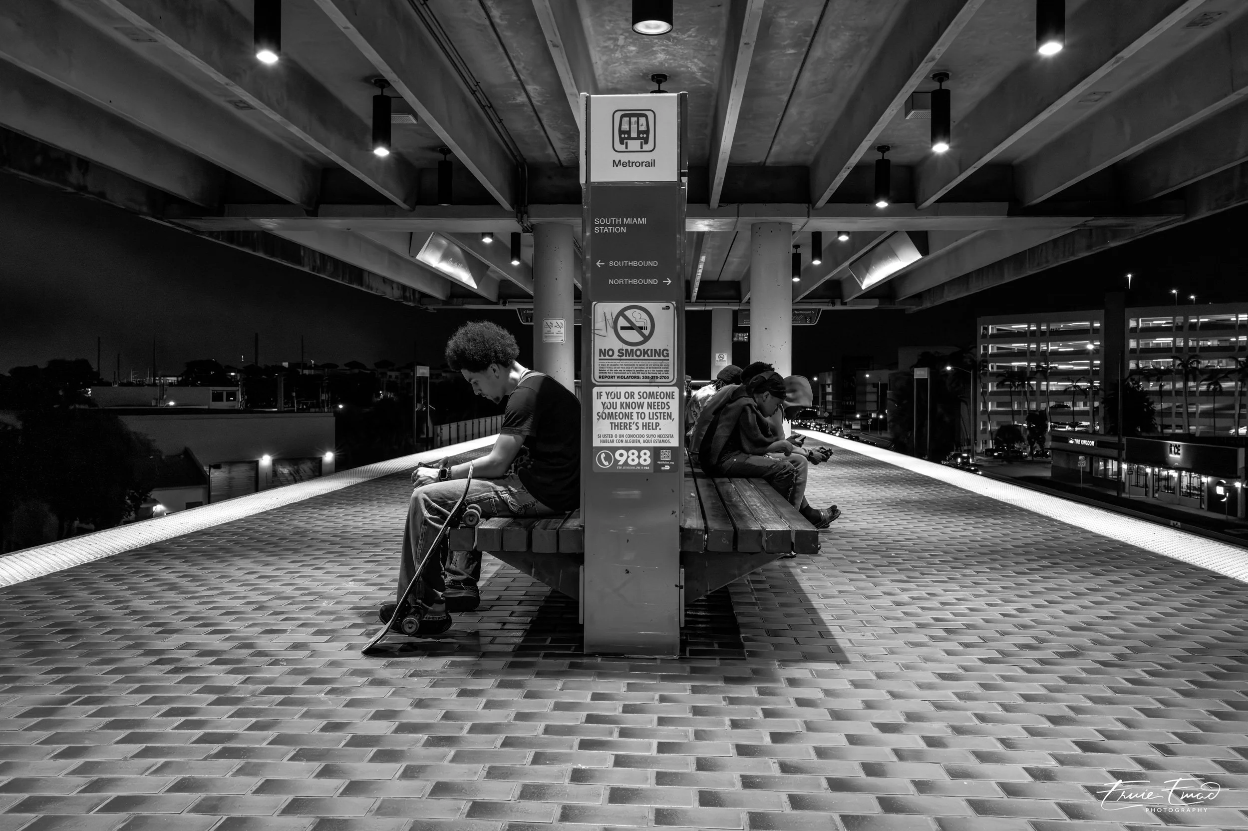 A black and white photo of a transit station platform at night with three people seated on a bench, one person with a skateboard, a sign in the middle indicating directions for the Metrorail and no smoking, and a cityscape with buildings and palm tre