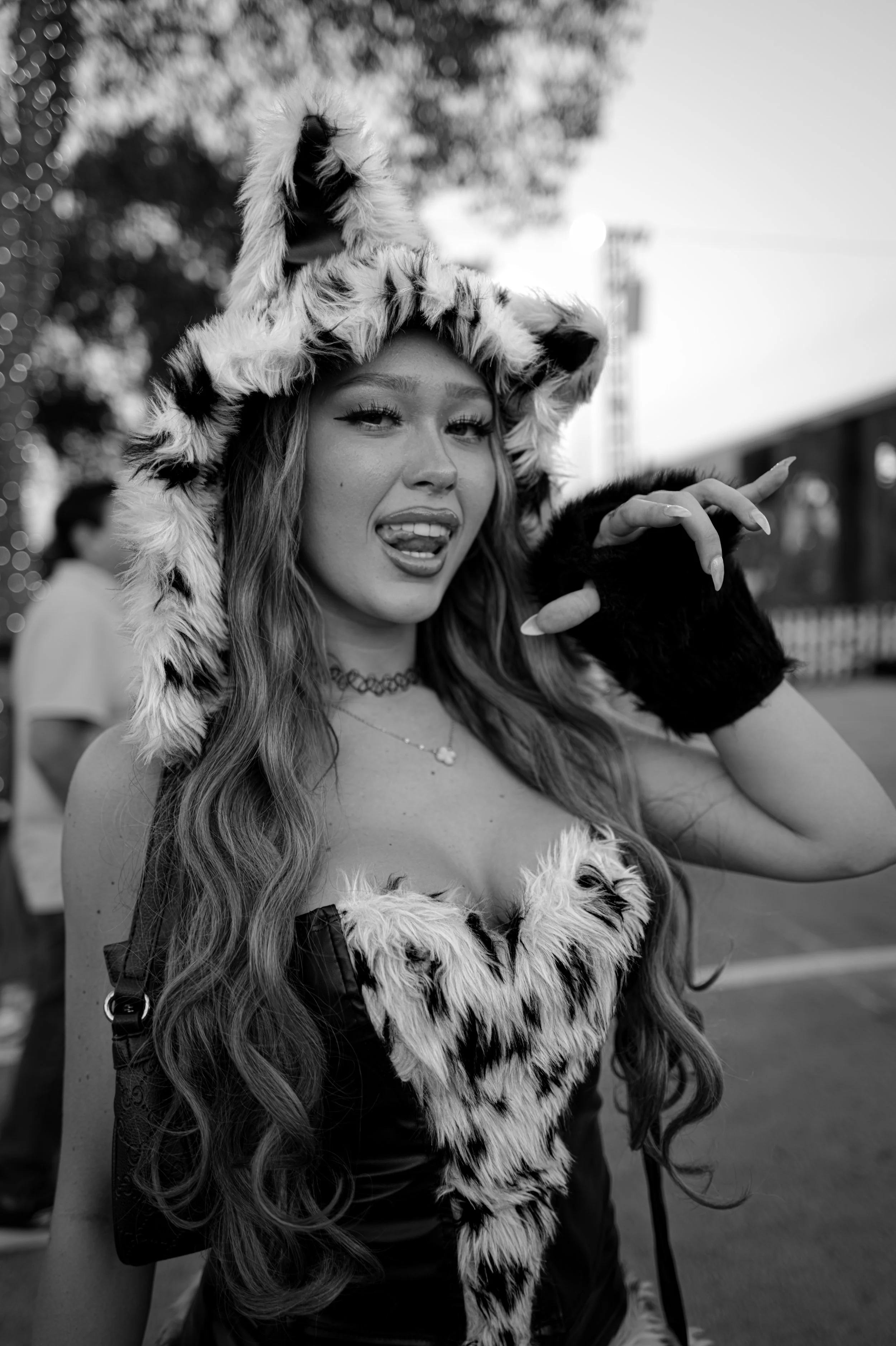 Woman wearing a furry animal costume top, making a playful expression with her tongue out and hand raised, outdoors during the daytime.