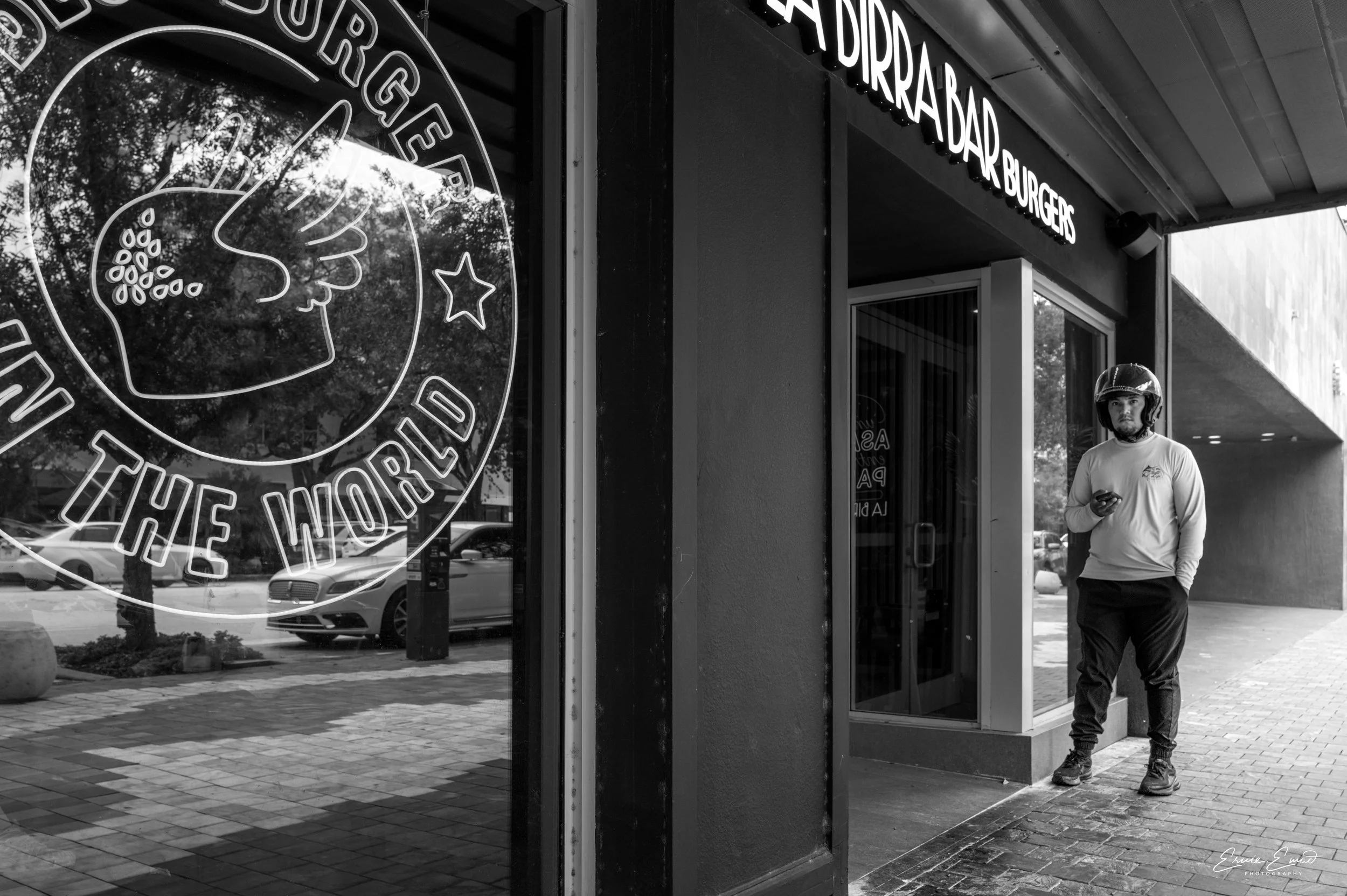 A man wearing a helmet and casual clothes is standing outside a burger restaurant, looking at his phone. The restaurant has large windows with signage, including a neon logo and the words 'BURGER BAR' on the exterior on Miracle Mile in Coral Gables