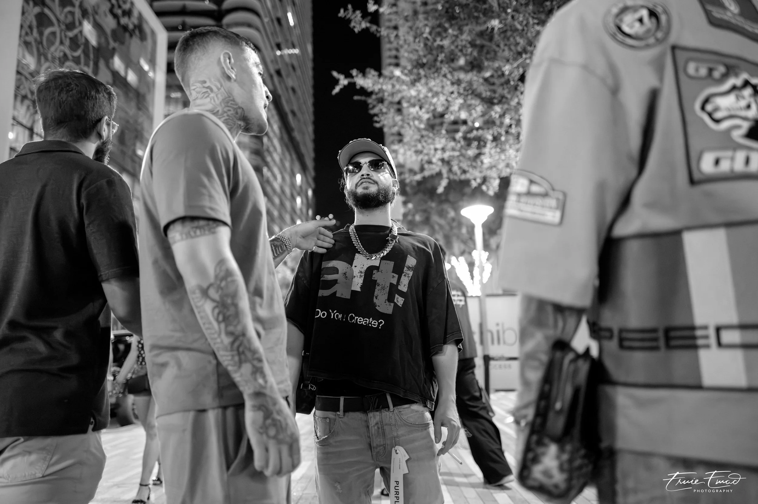 Four men engaged in conversation on a city street at night. One man is wearing sunglasses and a chain, while the others have tattoos and are dressed casually. They appear to be in a lively urban environment with buildings and lights in the background