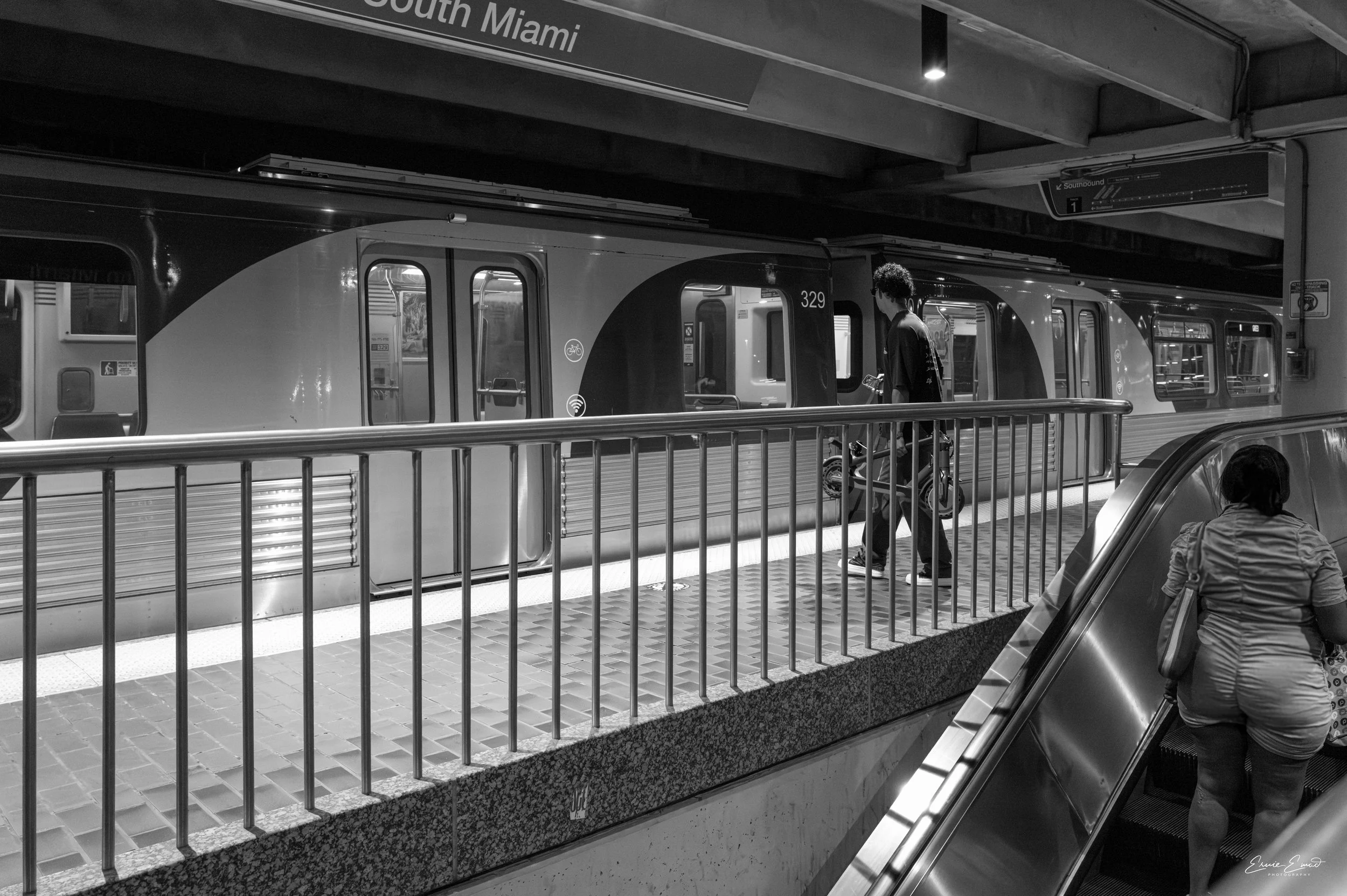 The Metrorail in South Miami with a skateboarder waiting for the doors to open from the arriving train while a lady takes the escalator to take the same train
