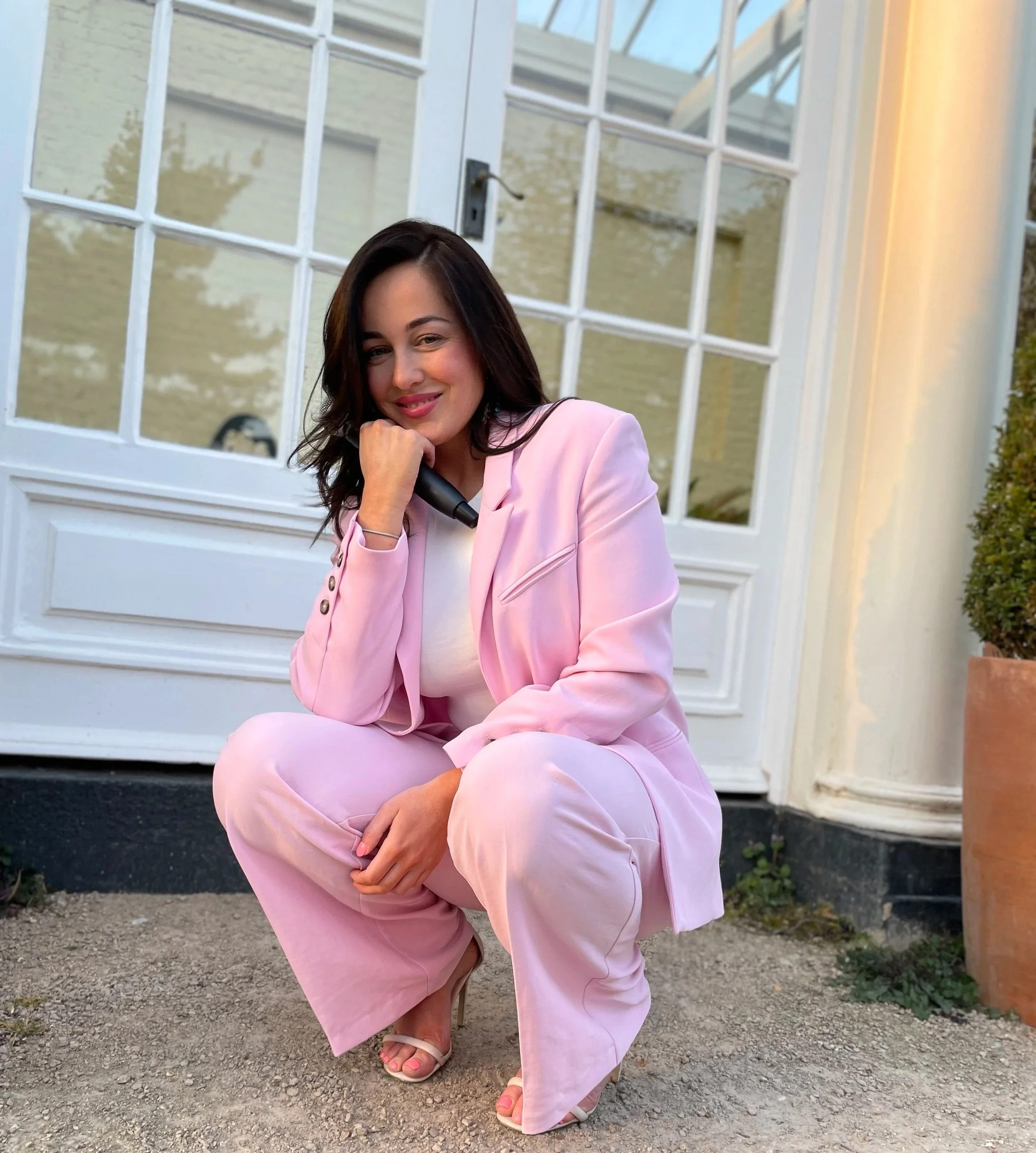 A woman with dark hair and fair skin dressed in a pink suit, squatting outdoors in front of a white building with glass windows, posing with her hand under her chin and smiling.