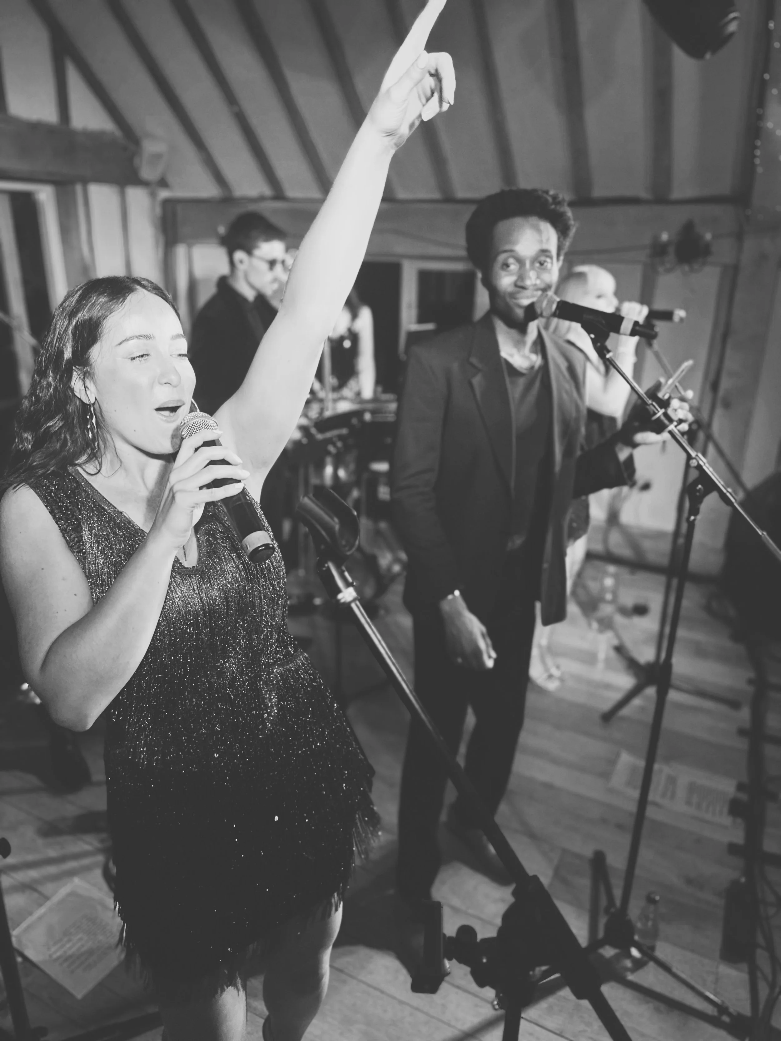A woman singing into a microphone with her eyes closed and her arm raised, while a man stands beside her holding a microphone and smiling, in a music or performance setting.