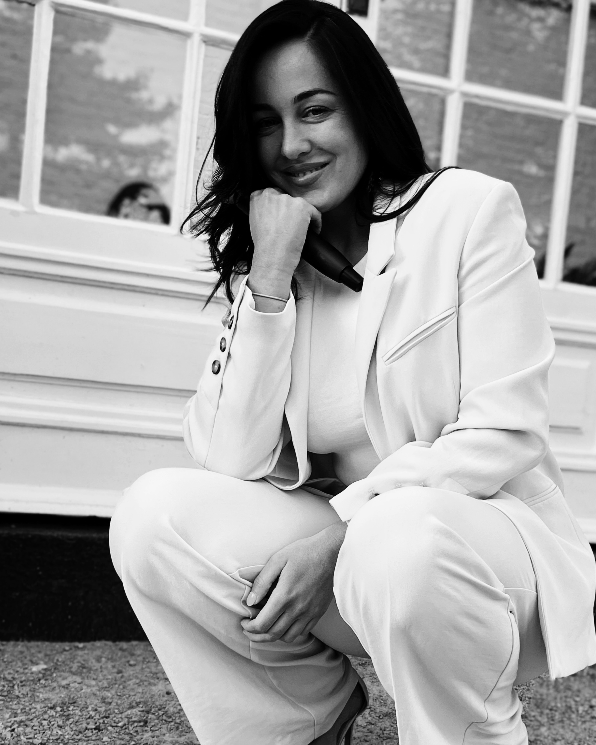 Woman with dark hair, wearing a white jacket, crouching outdoors, smiling with her chin resting on her hand, behind a windowed building.