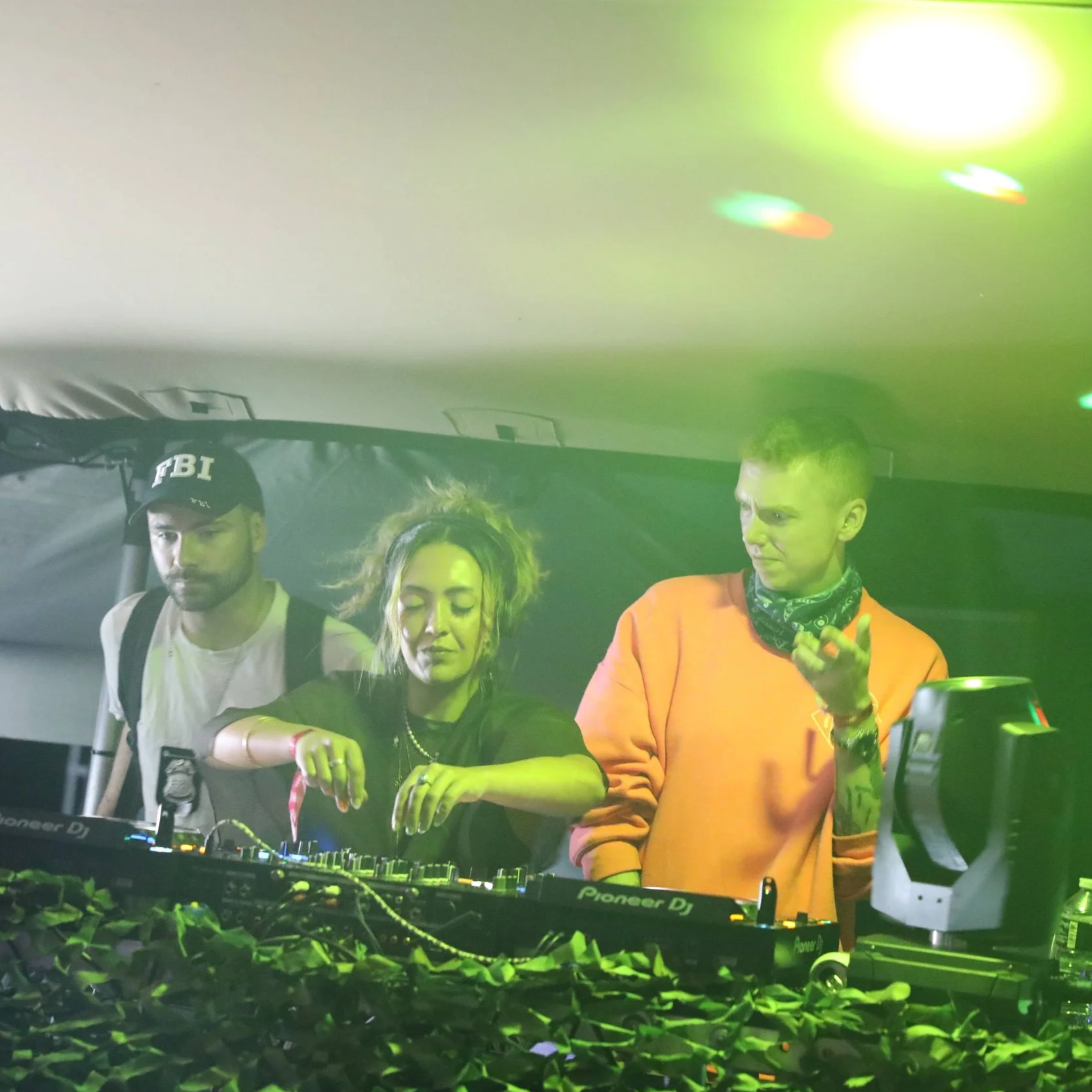 Three people at a DJ booth, with one man wearing an FBI hat, a woman adjusting equipment, and another man in an orange hoodie with a bandana around his neck, under green and yellow lighting.