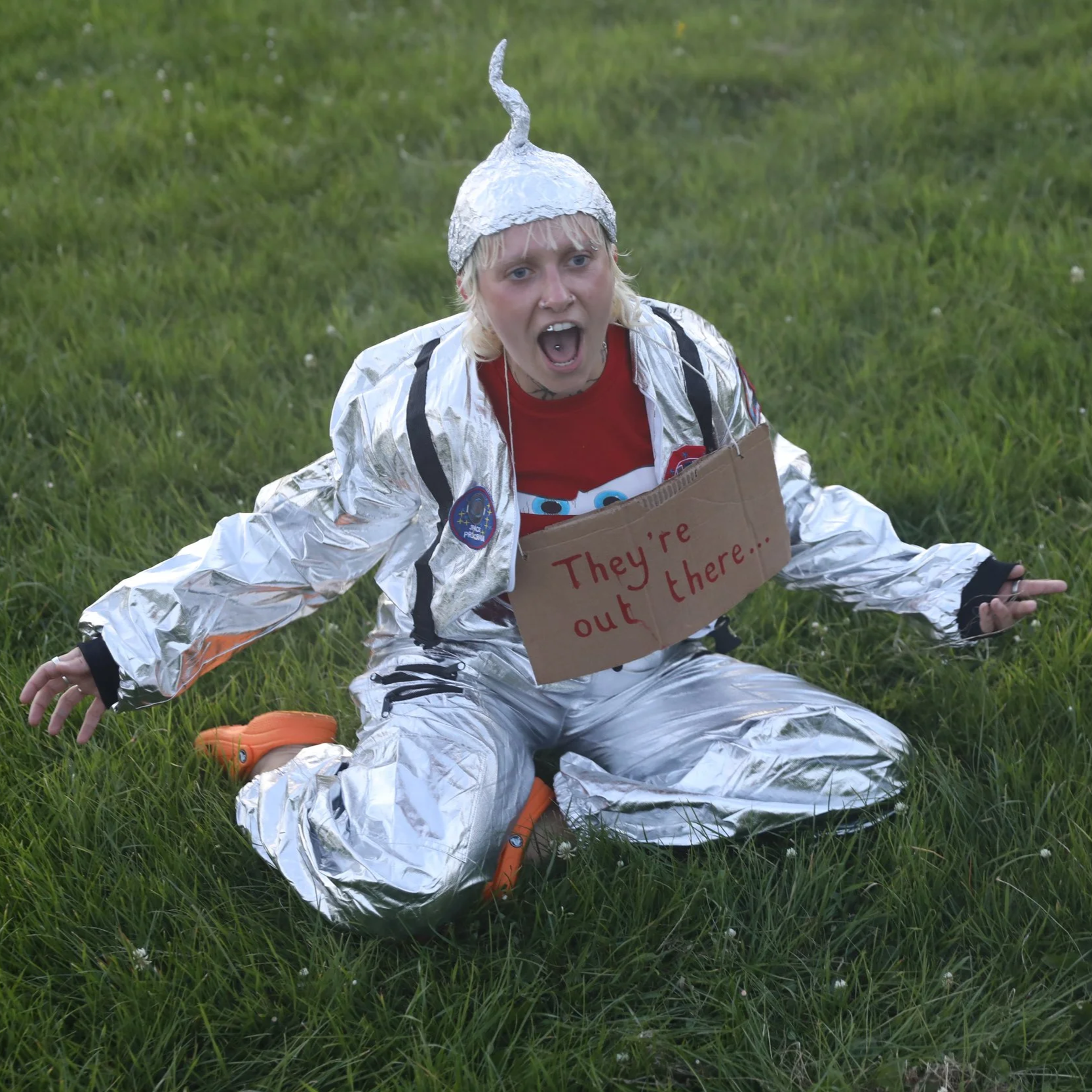 A person dressed as an astronaut in a shiny silver space suit, with a foil hat, is sitting on the grass with a surprised or shouting expression, and a cardboard sign that reads 'They're out there...' is around their neck.