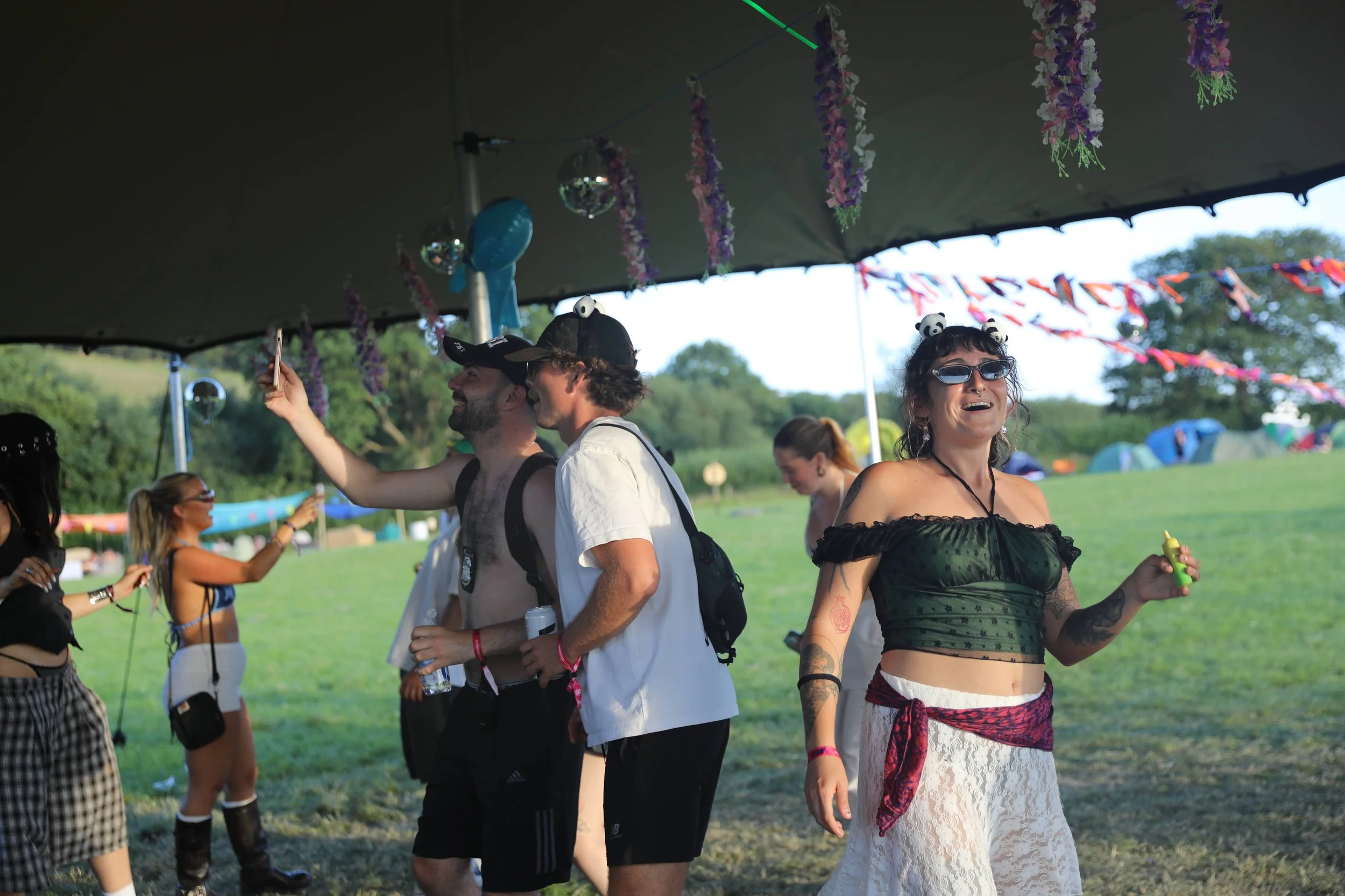 Group of people at an outdoor festival under a canopy, decorated with hanging flowers and disco balls, enjoying the event and taking photos.
