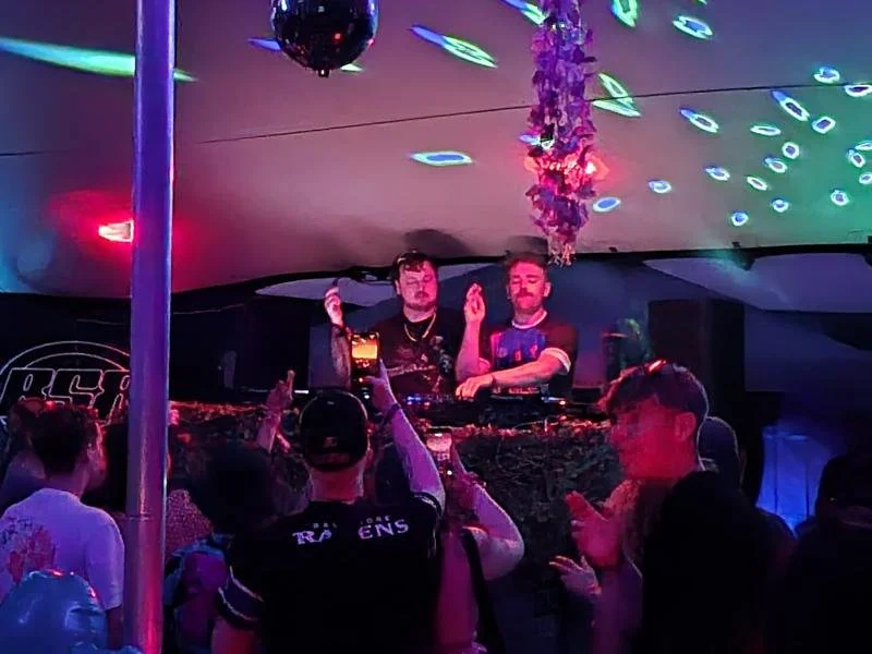 Two DJs perform on stage at a nightclub with colorful lighting, a disco ball, and a crowd dancing.