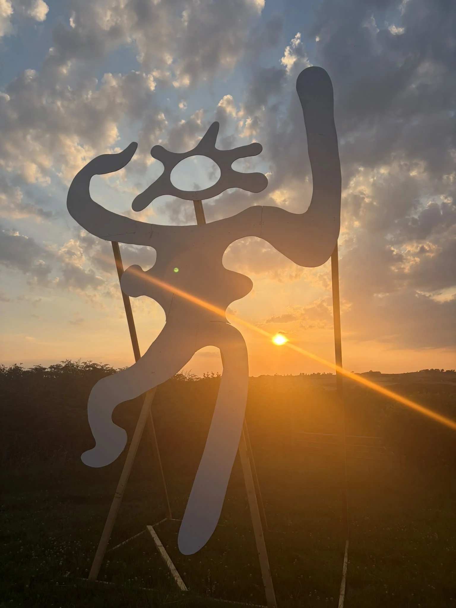 Silhouette of a cow-shaped cutout with a smaller cutout of a cow's head, set against a sunset sky with clouds and low horizon landscape.