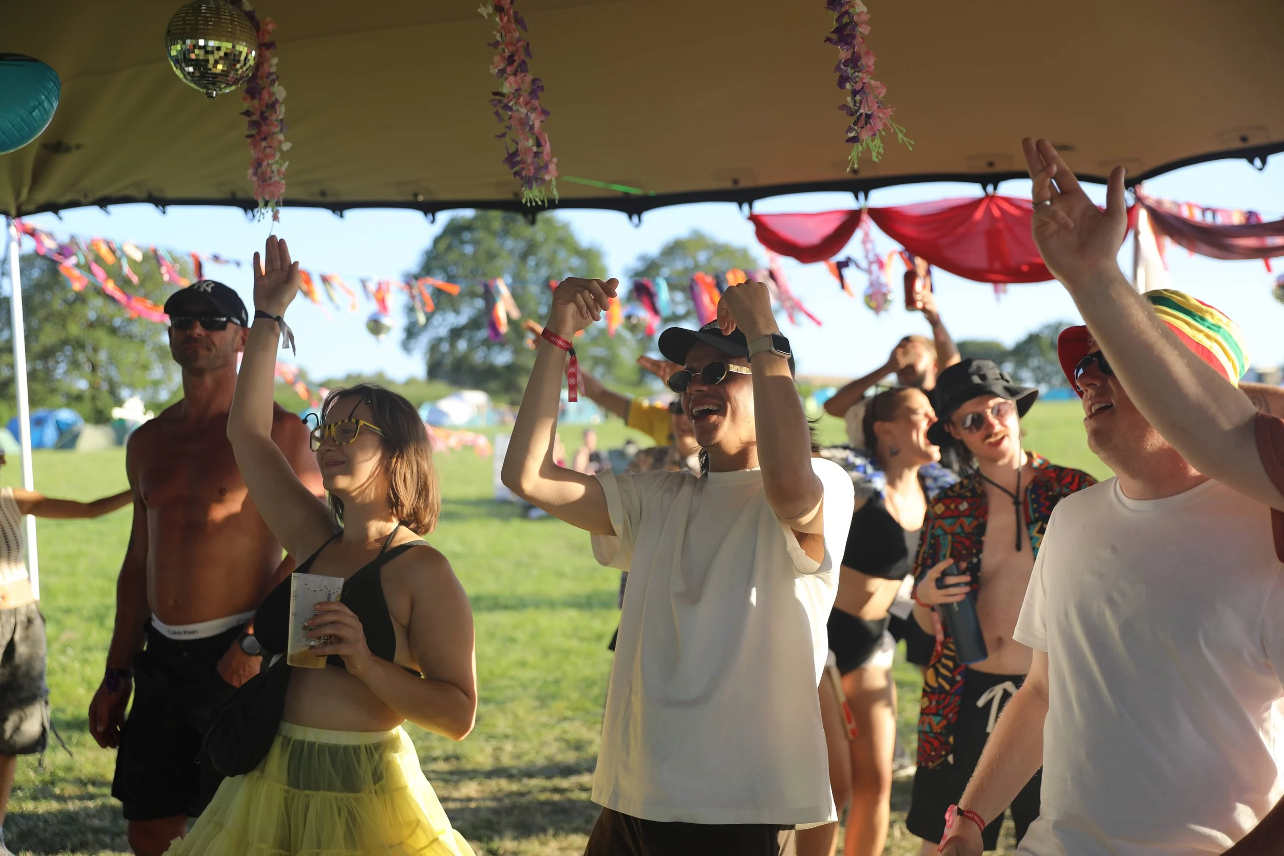 People dancing and smiling under a decorated canopy at an outdoor festival or party on a sunny day.