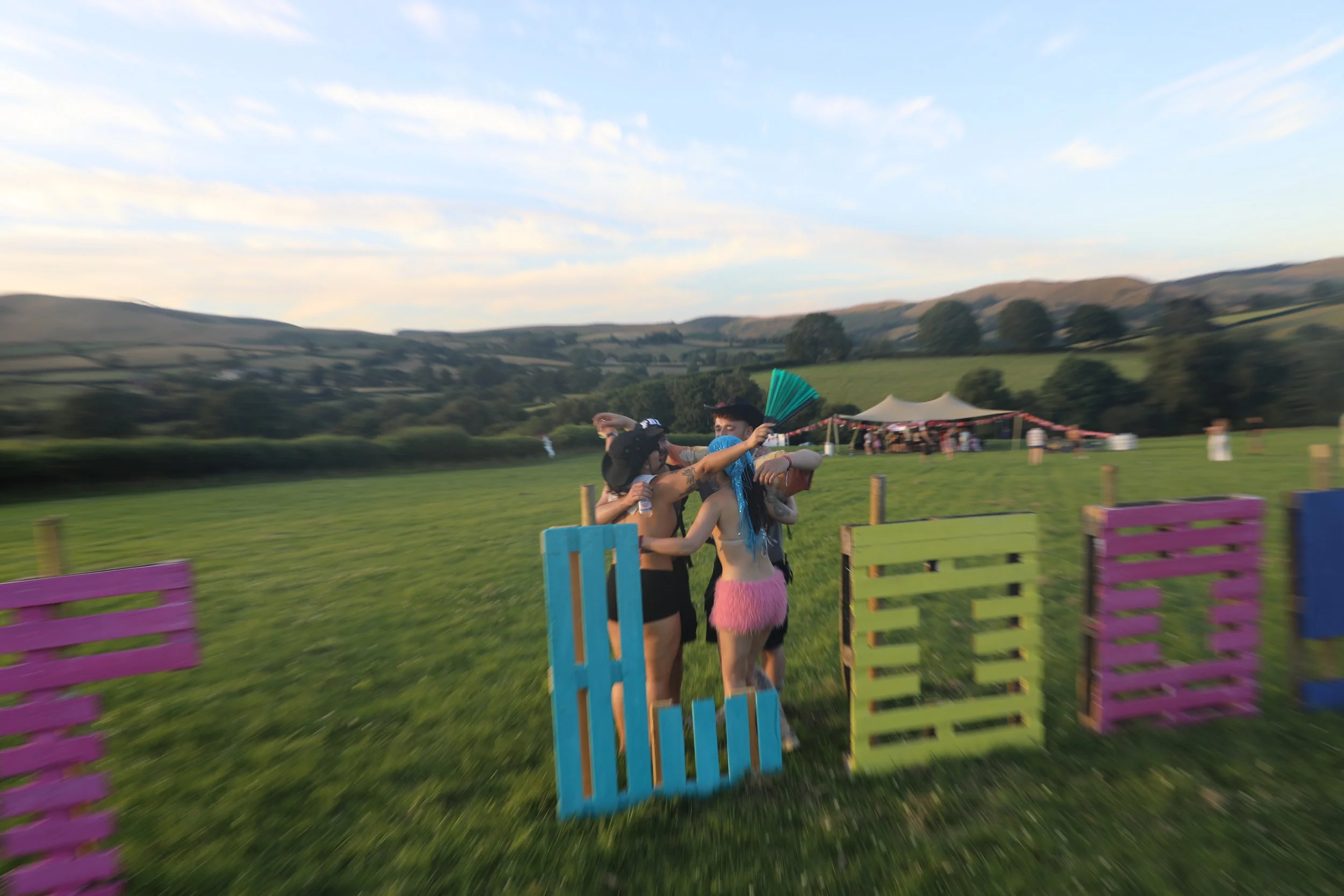 People enjoying a festival outdoors with colorful wooden barriers in a field, hills in the background, and a clear sky.