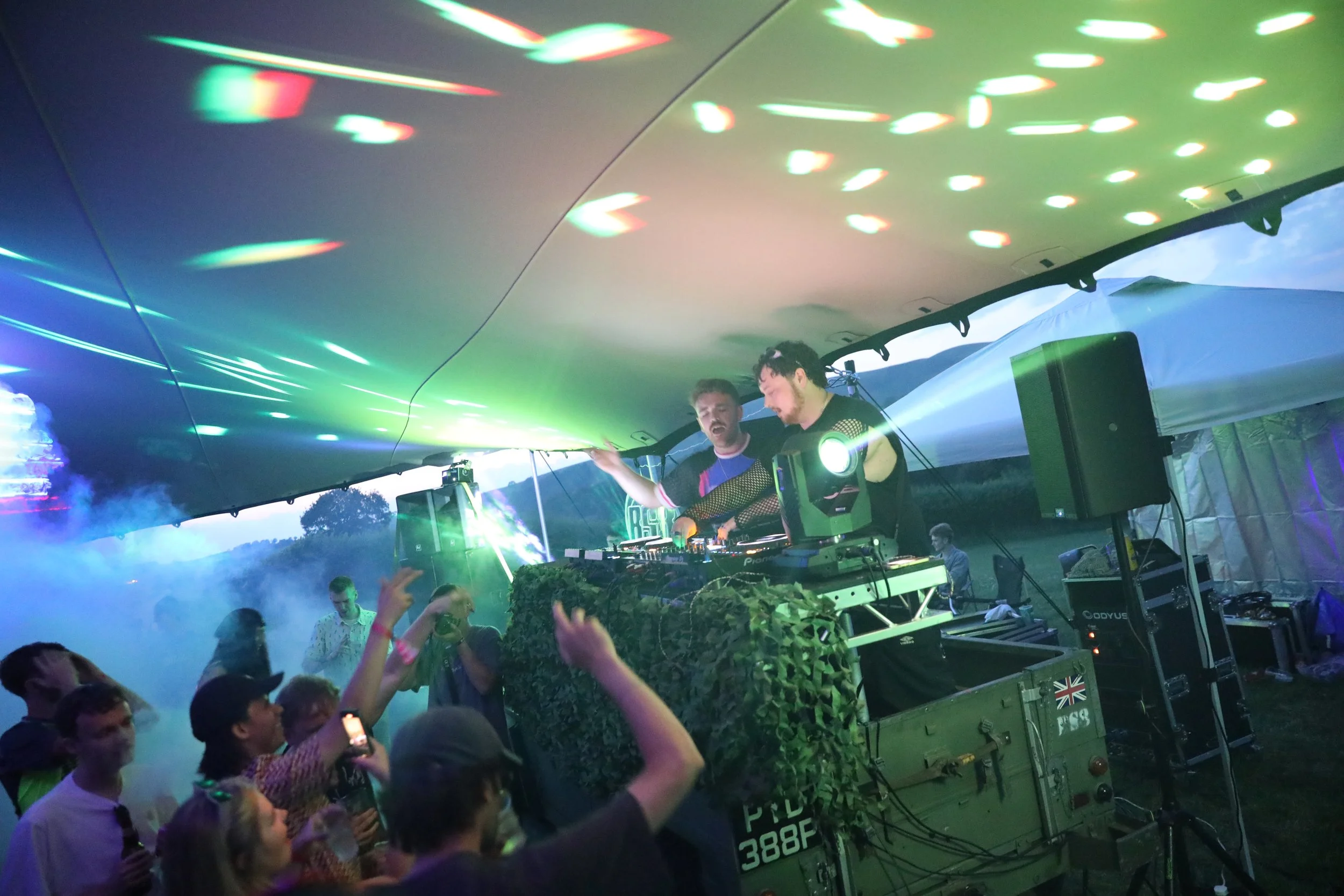 A DJ performs at an outdoor music event with colorful laser lights and a crowd dancing.
