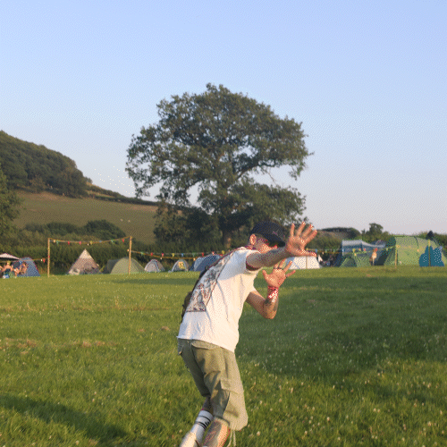 A person wearing a white shirt, khaki shorts, and sunglasses playing with a frisbee in a grassy park during daytime, with tents and trees in the background.
