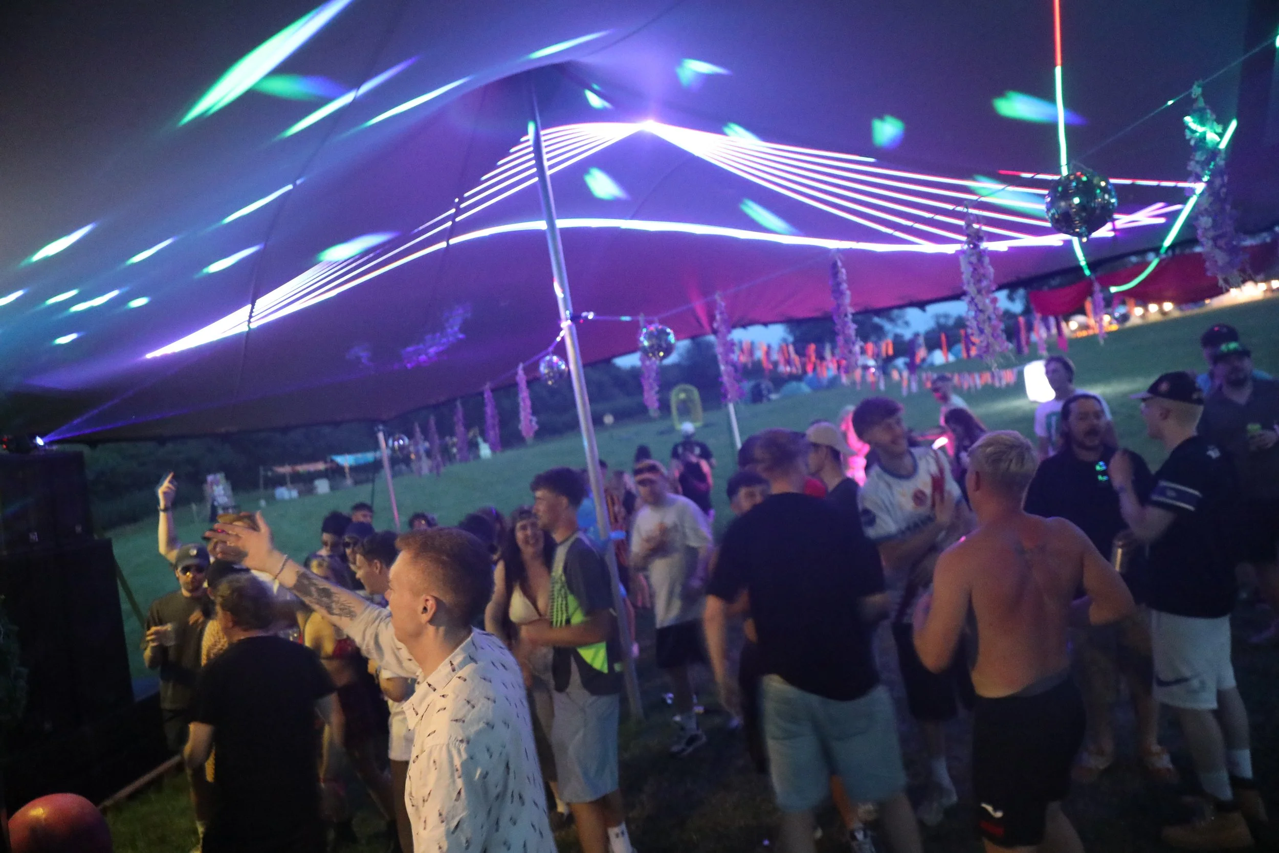People dancing and socializing at an outdoor night event with colorful laser light displays and hanging disco balls.