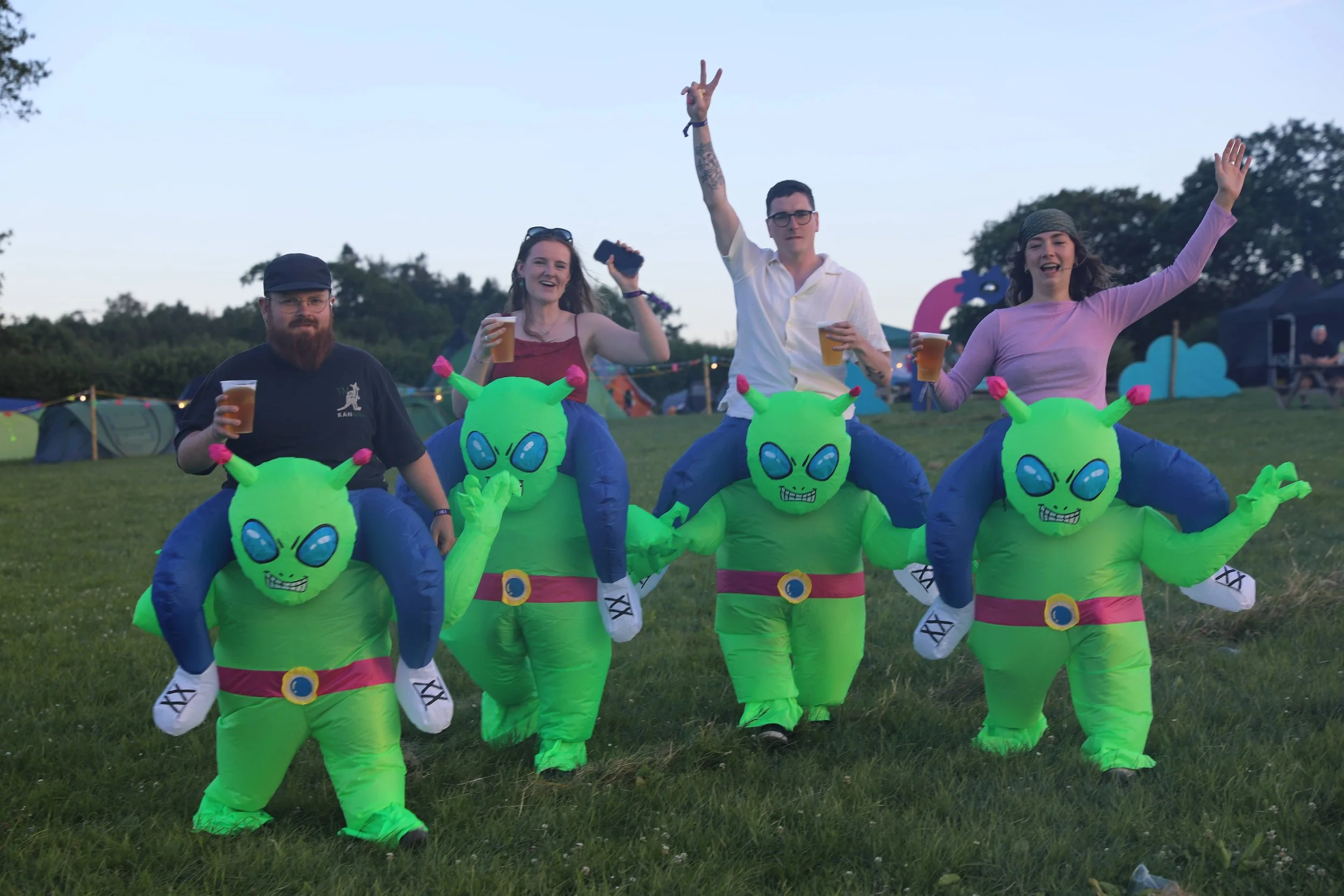 People wearing inflatable alien costumes and holding drinks at an outdoor event in a field during evening.