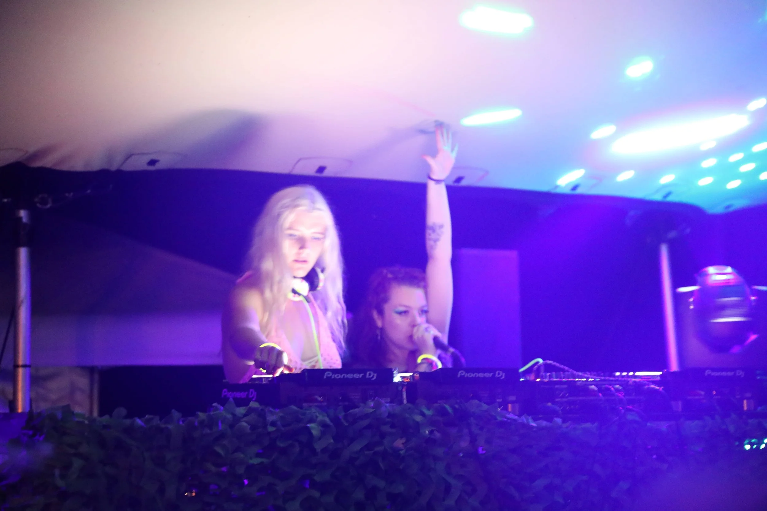 Two female DJs performing at a nightclub or party, illuminated by colorful purple, pink, and blue lights, with DJ equipment in front of them.