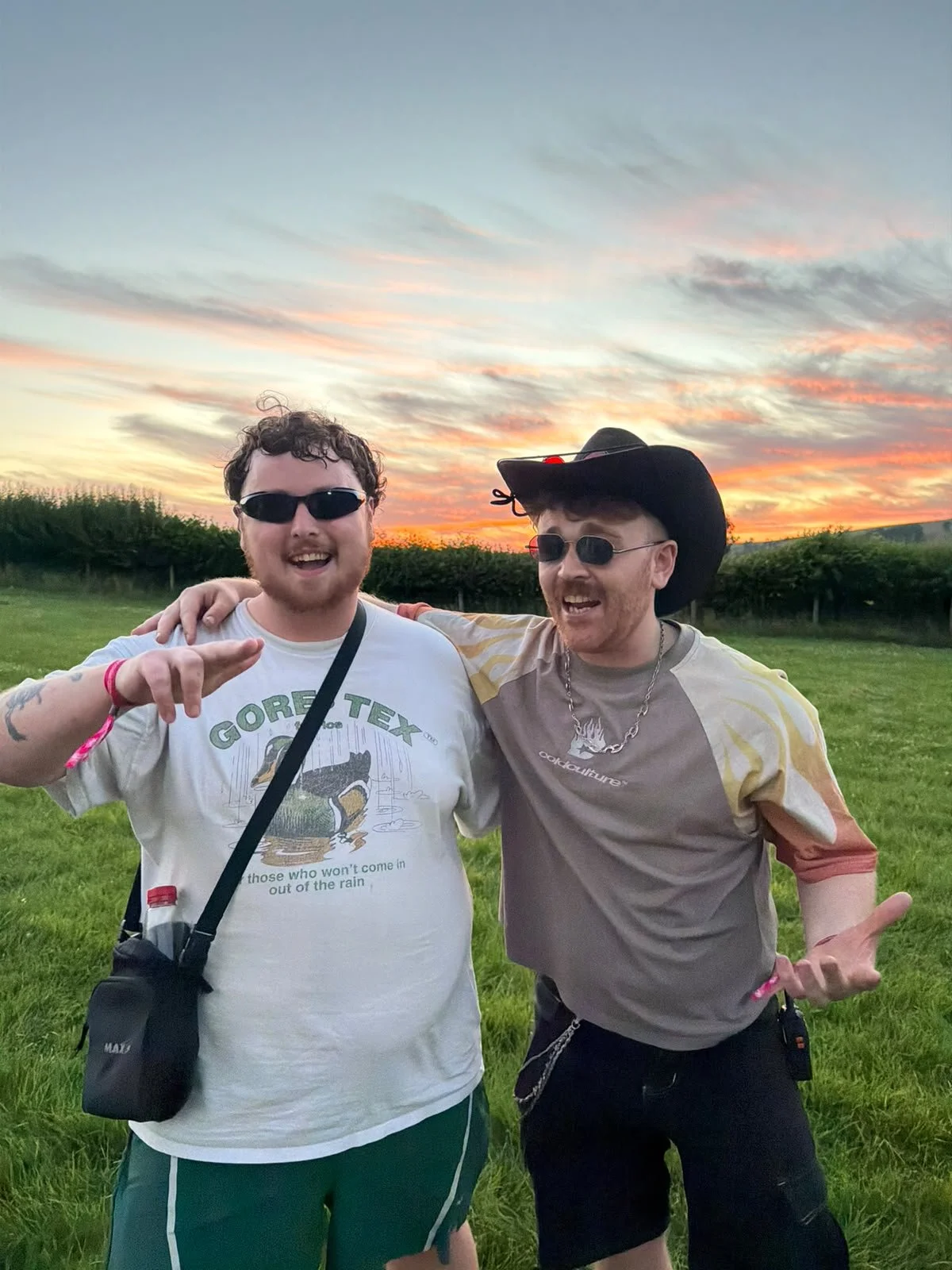 Two men smiling and posing outdoors during sunset. They are wearing sunglasses and casual clothes, one with a cowboy hat, with their arms around each other in a grassy field with a sunset sky in the background.