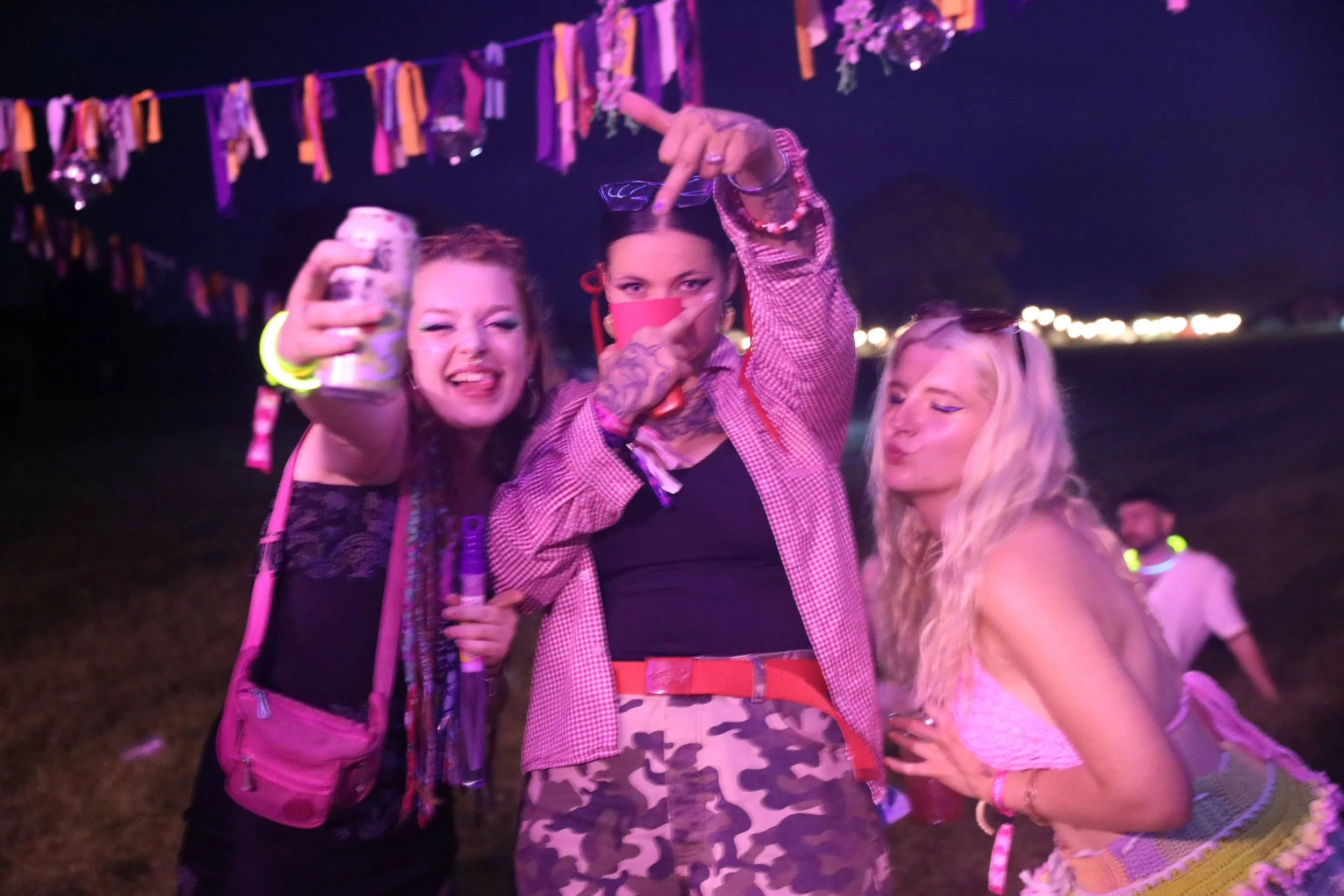 Three women celebrating at night outdoors, with colorful decorations hanging in the background. One woman is holding a drink and making a gesture with her hand, another woman is holding a beverage, and the third woman is leaning in close, smiling.