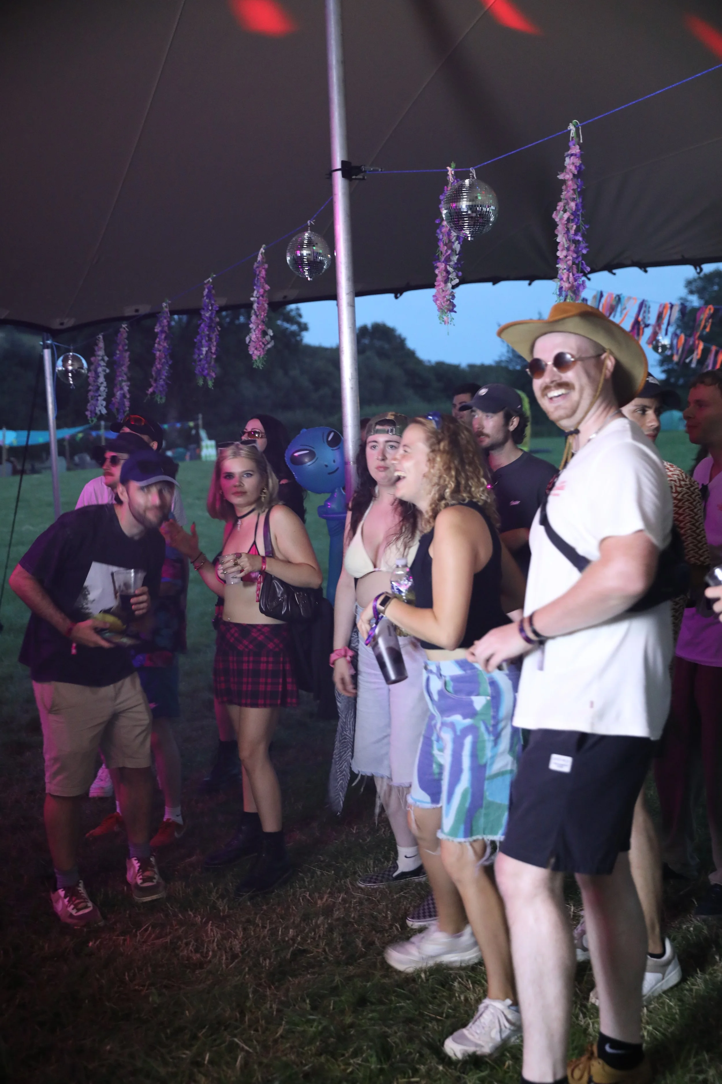 A group of young people at an outdoor event under a tent, decorated with hanging purple flowers and disco balls, with some holding drinks and smiling, dressed casually in summer clothing.