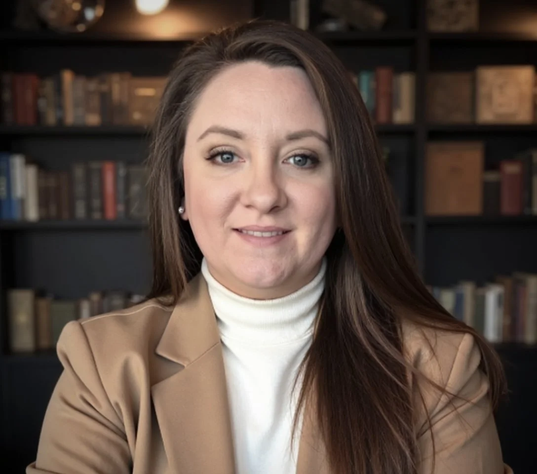 A woman with long brown hair, blue eyes, wearing a tan blazer over a white turtleneck, seated in front of a bookshelf filled with books.