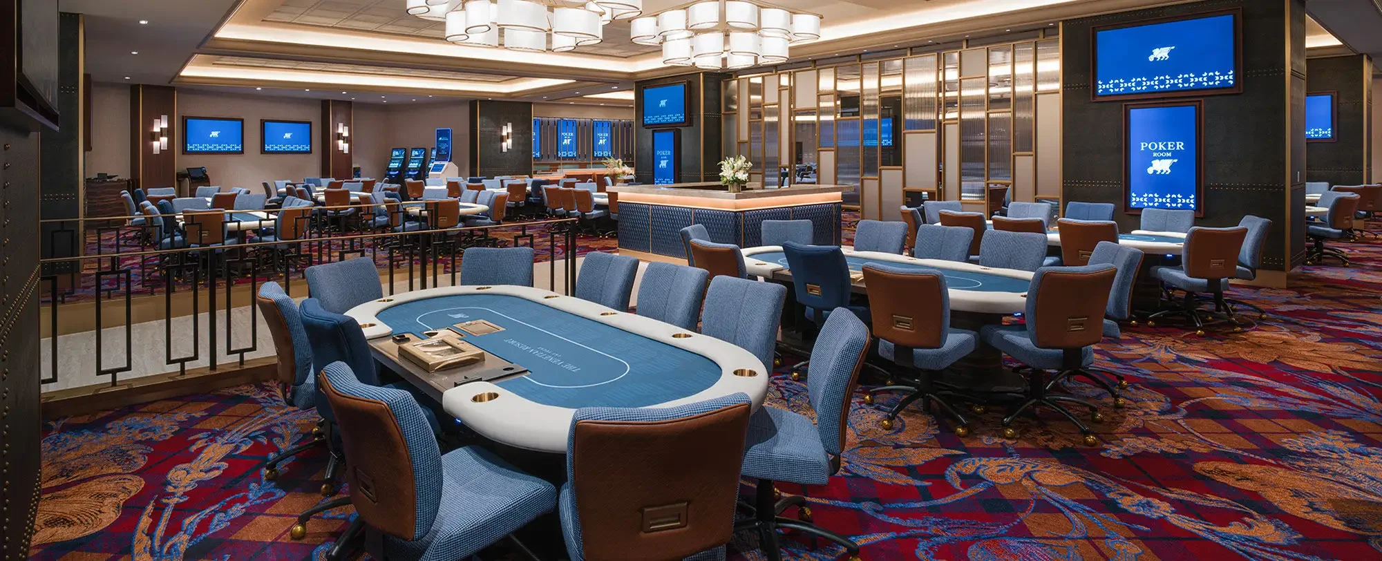 Casino poker room with blue poker tables, chairs, and digital screens, carpeted floor, and ceiling lights.