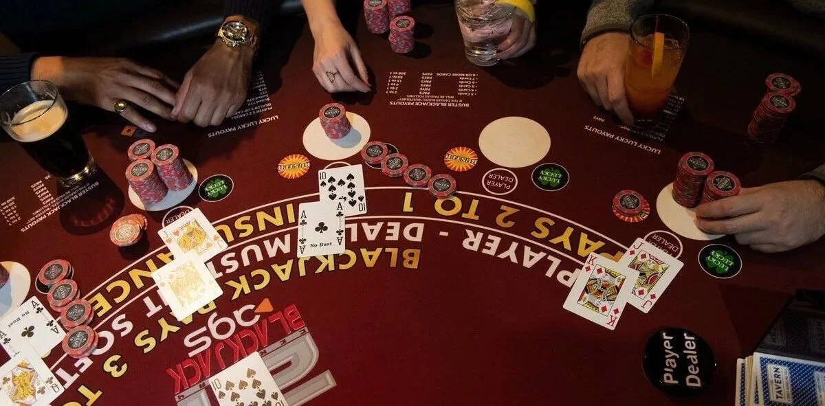 A poker game in progress with chips, playing cards, drinks, and personal items on a felt table. Four players are involved, seen with hands and drinks.