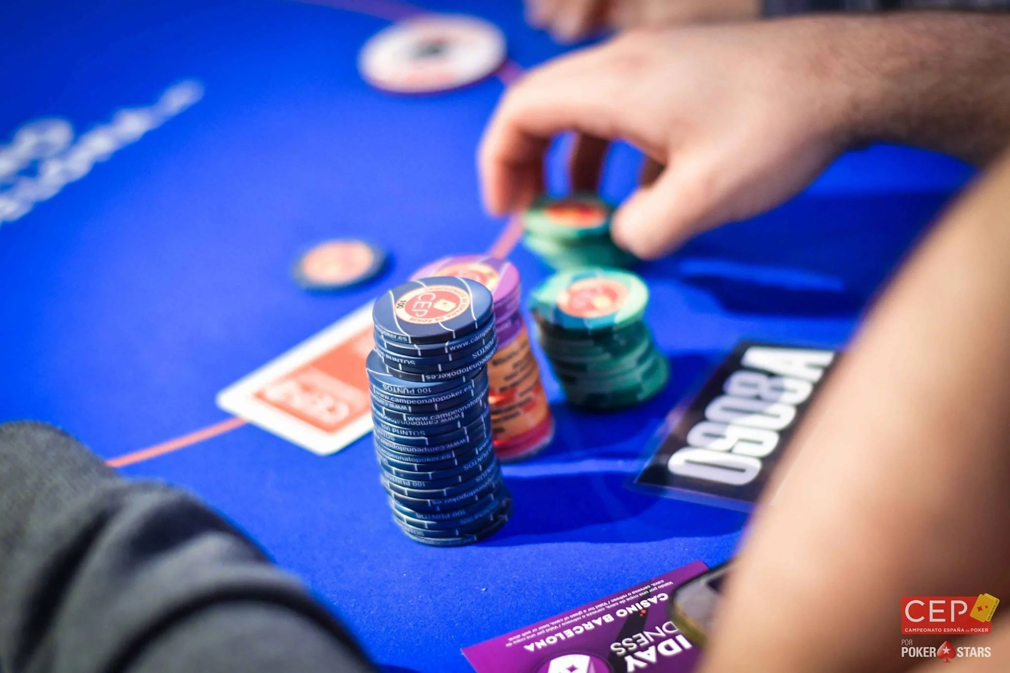 Close-up of poker chips stacked on a blue poker table during a game, with some promotional material on the table and part of the player's arm visible.