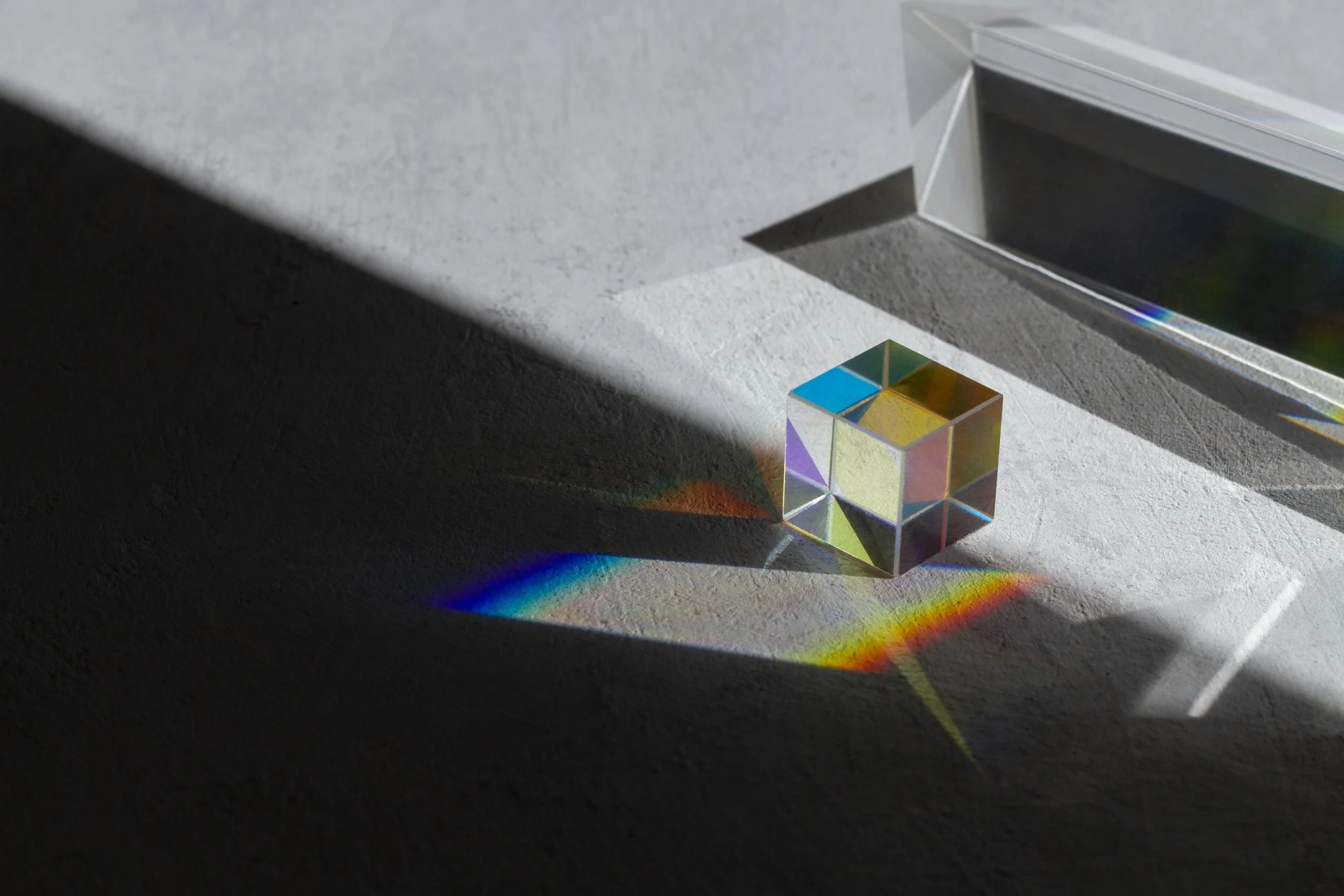 Cubos de acrílico transparente refletindo luz, projetando um arco-íris no piso de cimento de cor cinza escuro, com uma janela de alumínio ao fundo.