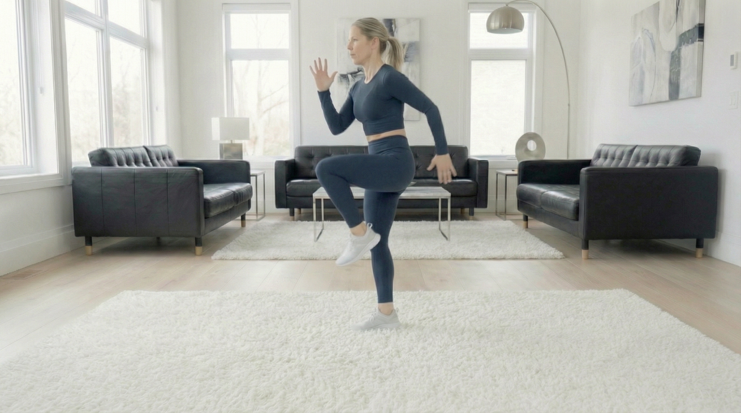 A woman with blonde hair in a ponytail wearing a dark blue workout outfit and white sneakers is exercising in a bright living room with large windows, black sofas, and modern decor.