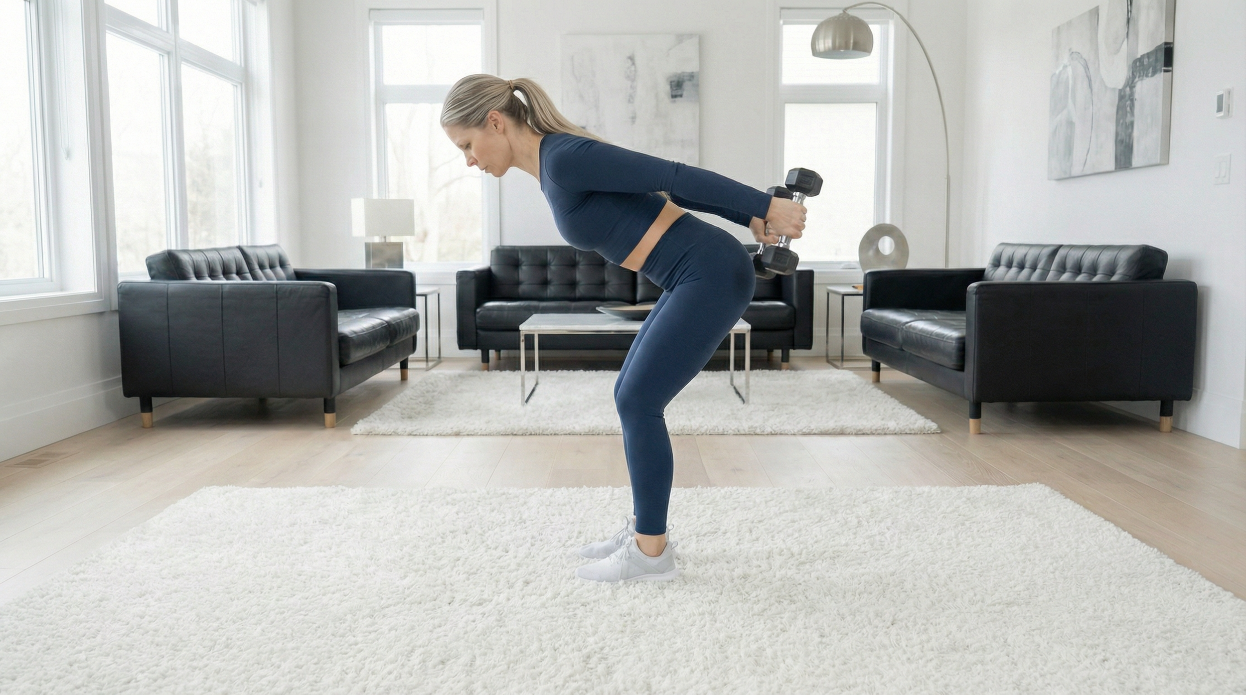 A woman exercising at home, lifting a dumbbell behind her back, on a white shag rug in a bright living room with black sofas, large windows, and modern artwork.