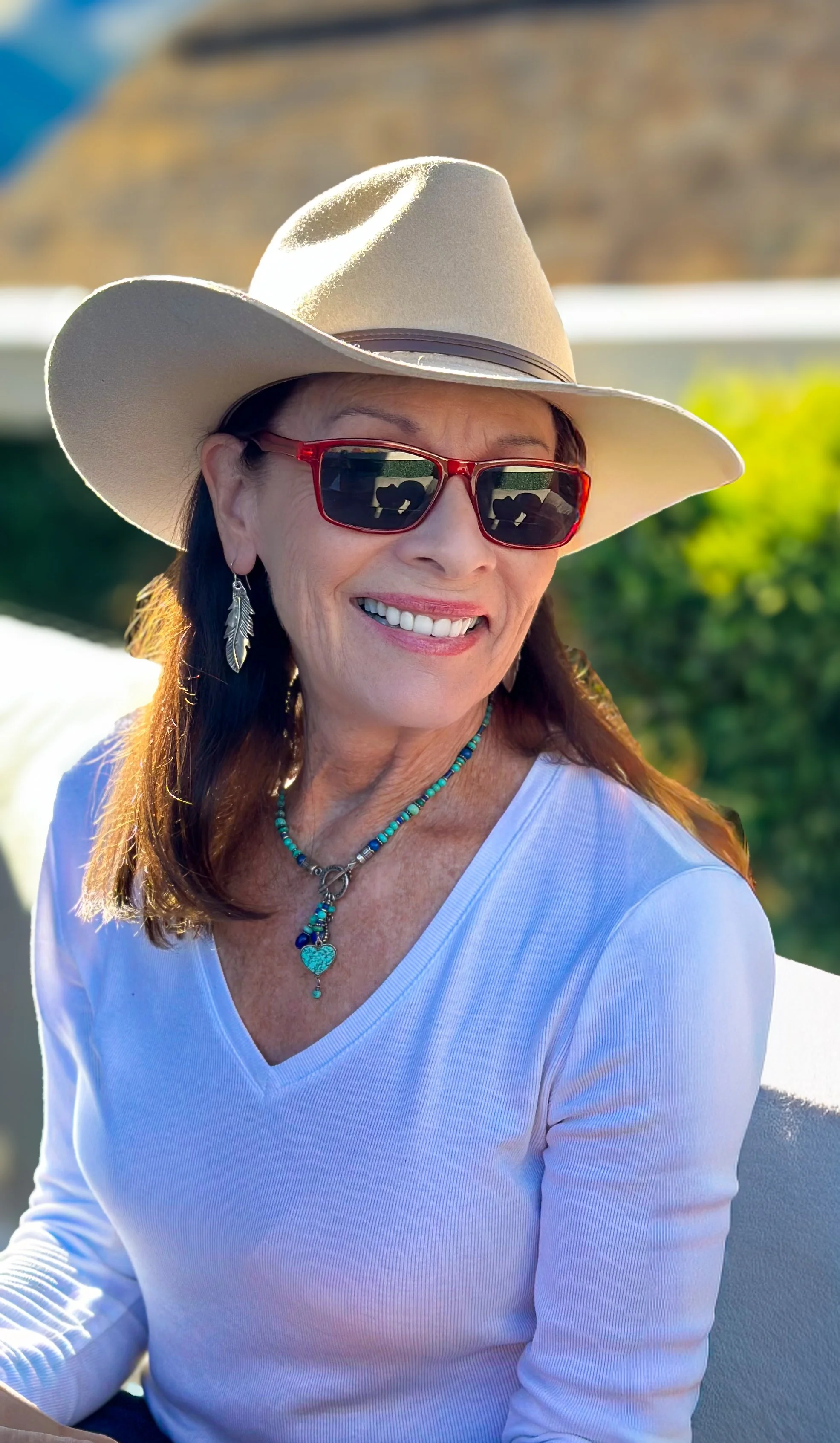 A smiling woman wearing a white wide-brimmed hat, red sunglasses, turquoise jewelry, and a white shirt outdoors on a sunny day.
