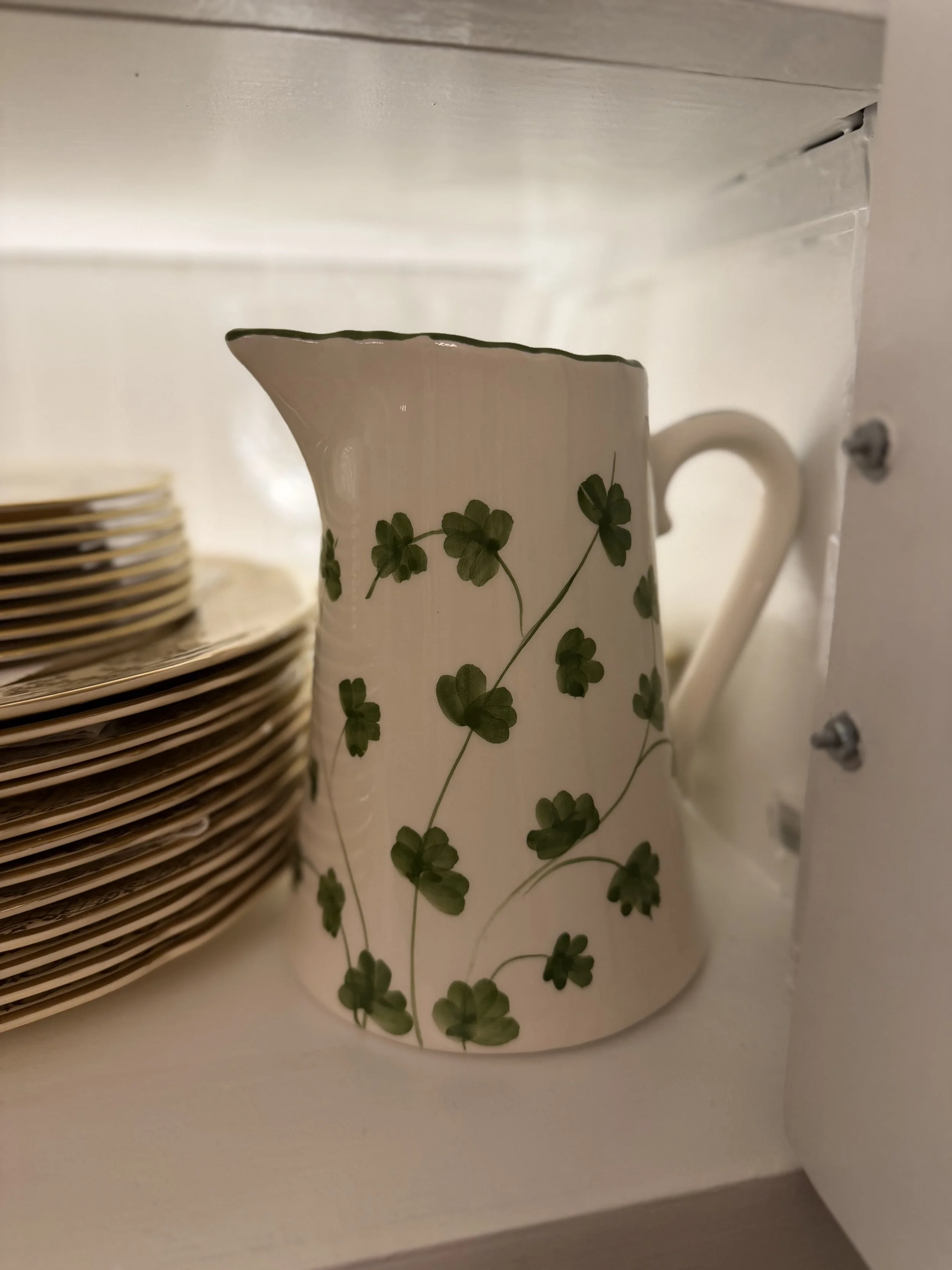 White ceramic pitcher decorated with green clovers beside stacked dishes in the dining room hutch