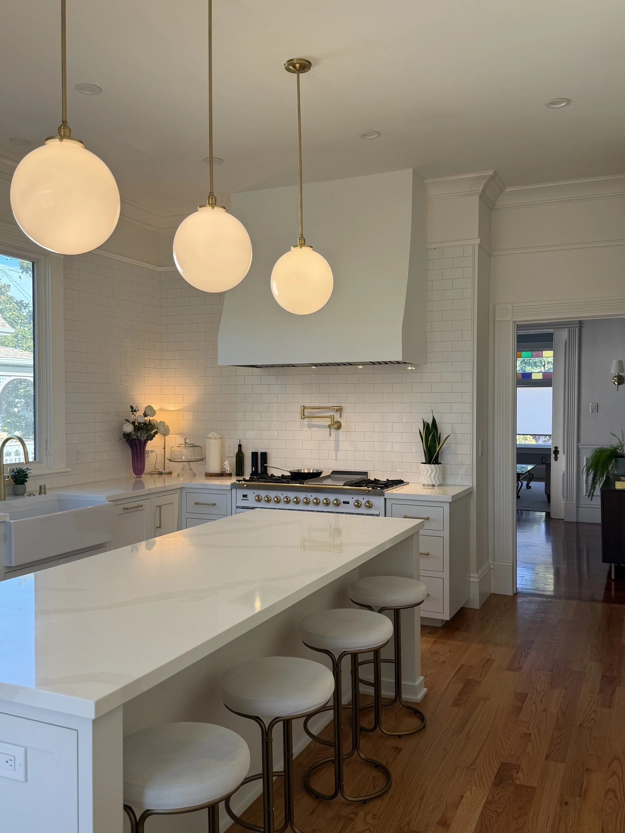 White kitchen in a 1900 house with a large range, statement hood, brass fixtures and three hand-blown glass pendant lights.