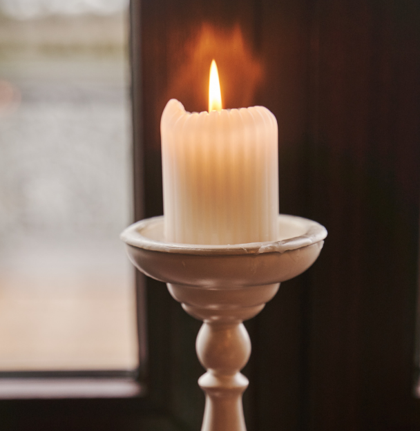 Lit cream pillar candle in a wooden candlestick beside a window