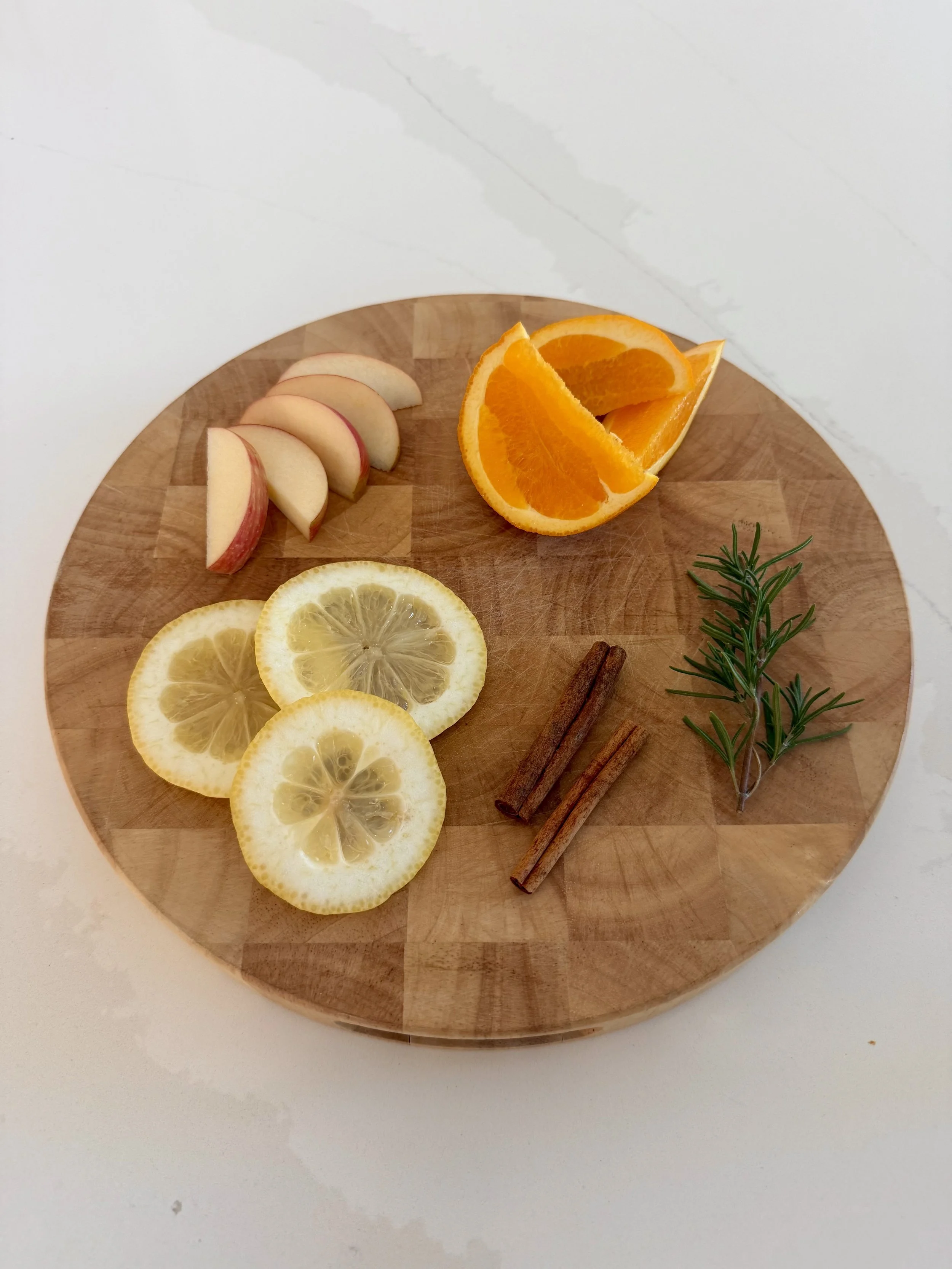 Fresh rosemary, lemon slices, orange wedges, apple slices and cinnamon sticks arranged on a round wooden cutting board
