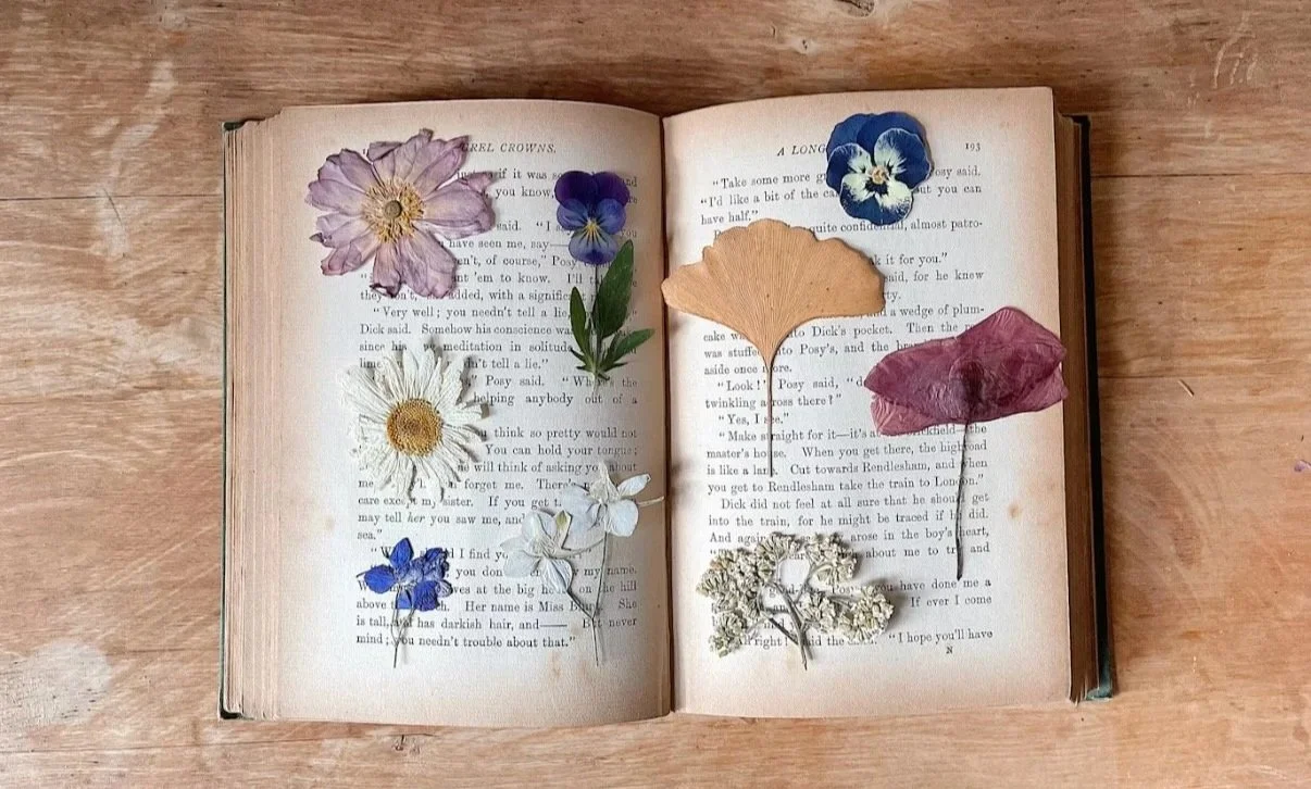 An open vintage book, resting on a warm wooden surface, with an assortment of colorful pressed flowers in soft pink, purple, blue, and golden tones arranged across its pages