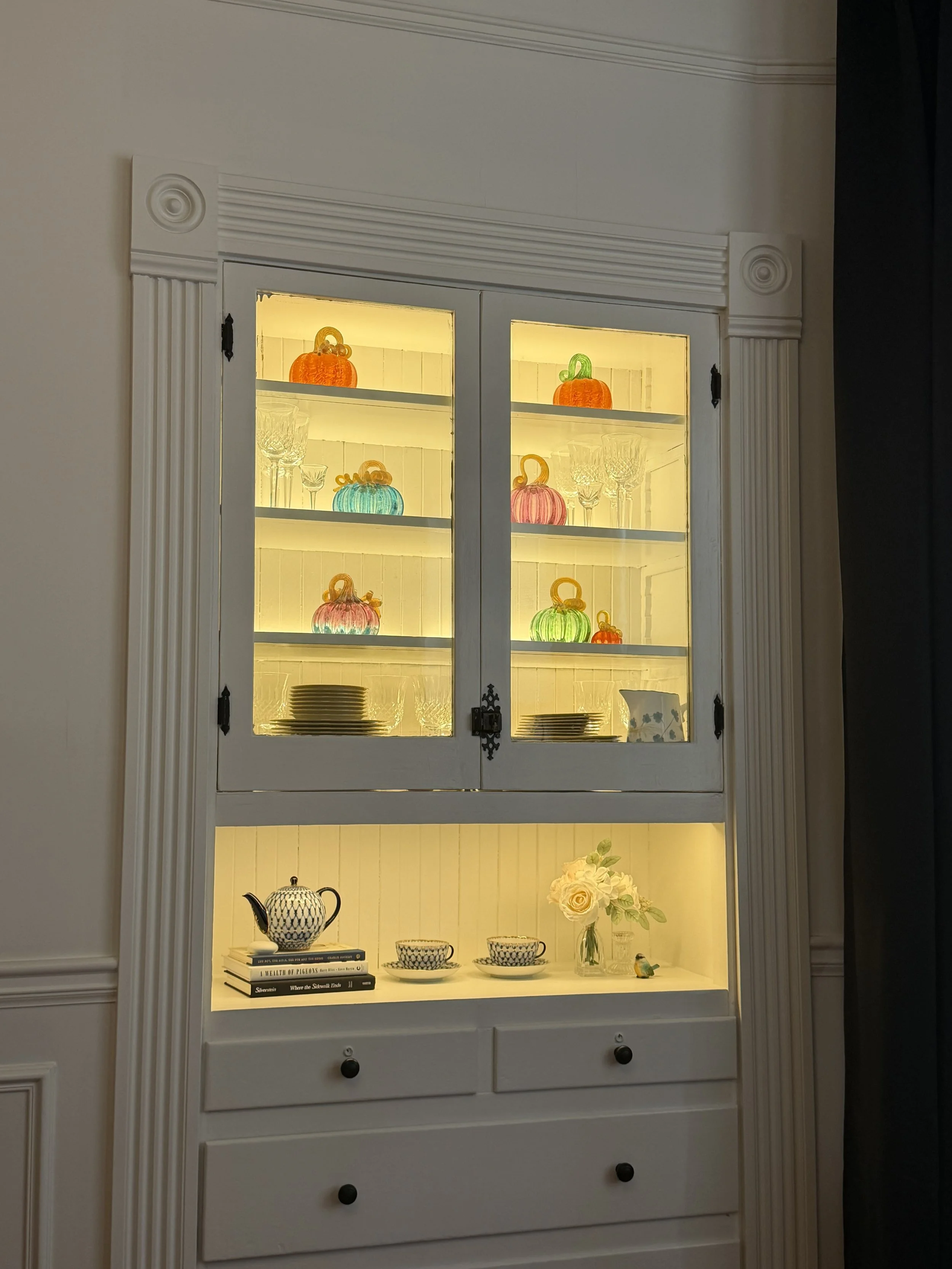 Built-in dining room hutch glowing in the evening with colorful glass pumpkins, dishes and flowers