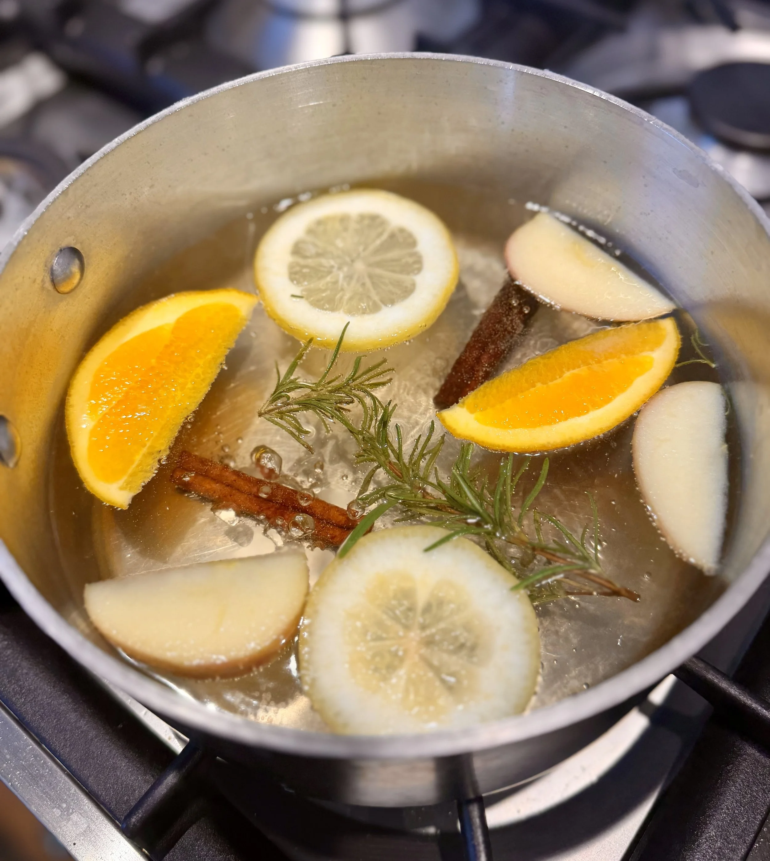 Fresh rosemary, orange wedges, lemon slices, apple slices and cinnamon sticks in a simmer pot on the stove