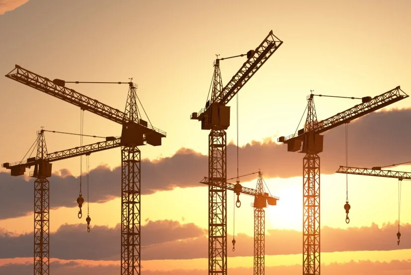 Cinq grues de construction silhouettées contre un ciel au coucher du soleil.